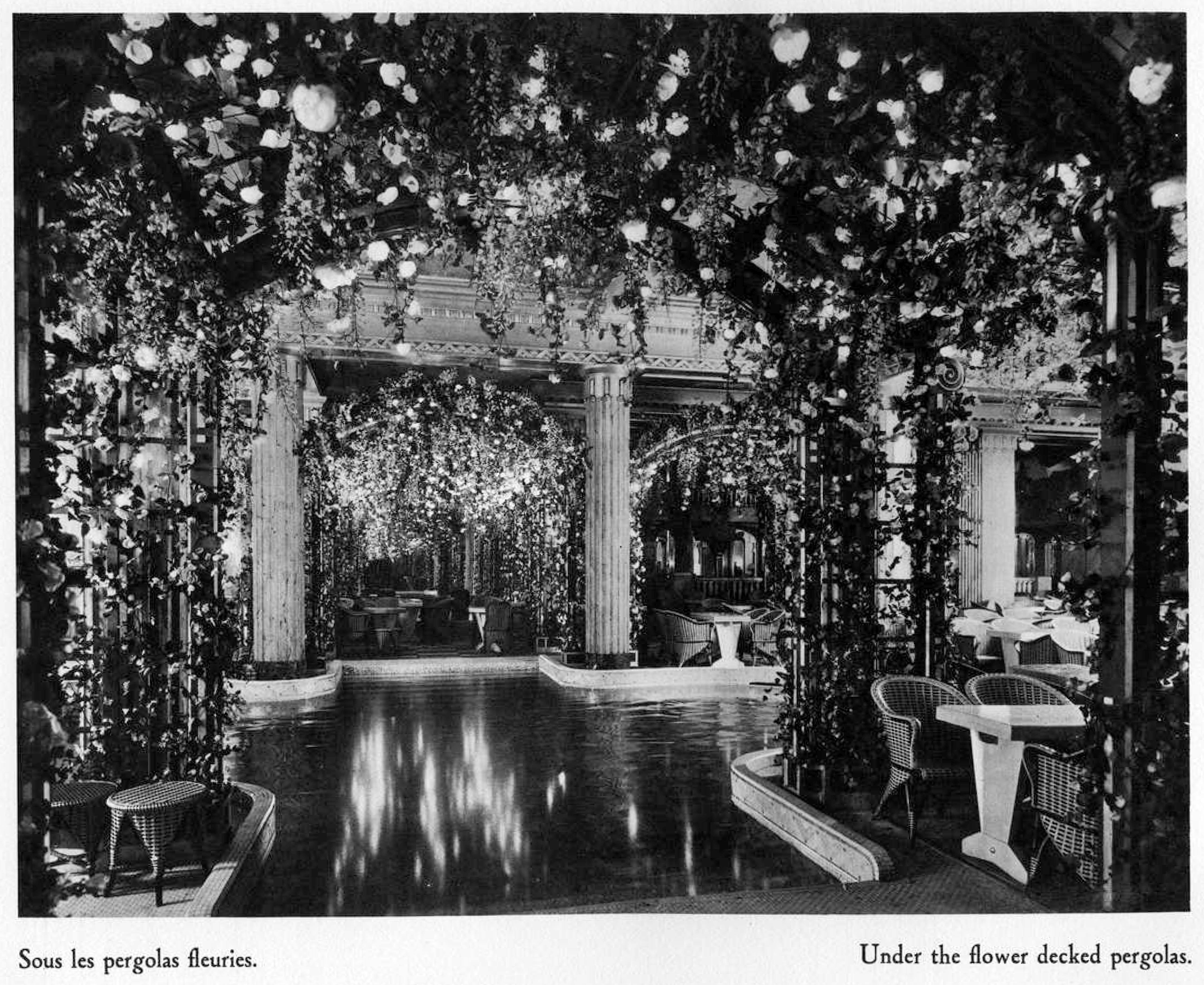 Black and white image of an interior pool surrounded by seating and covered by flowered pergolas