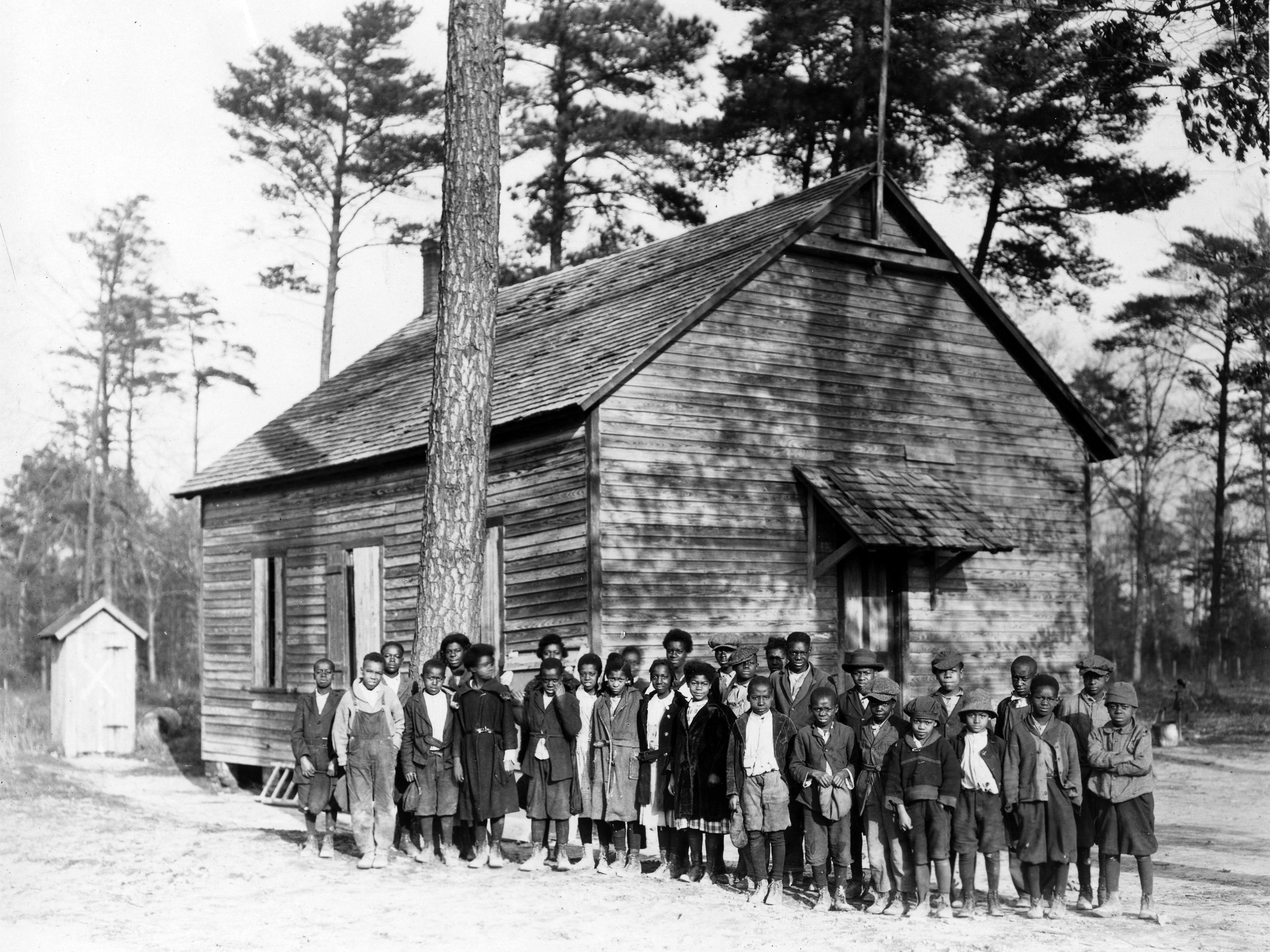 Black and white photo of young, Black schoolchildren outside a one room schoolhouse.