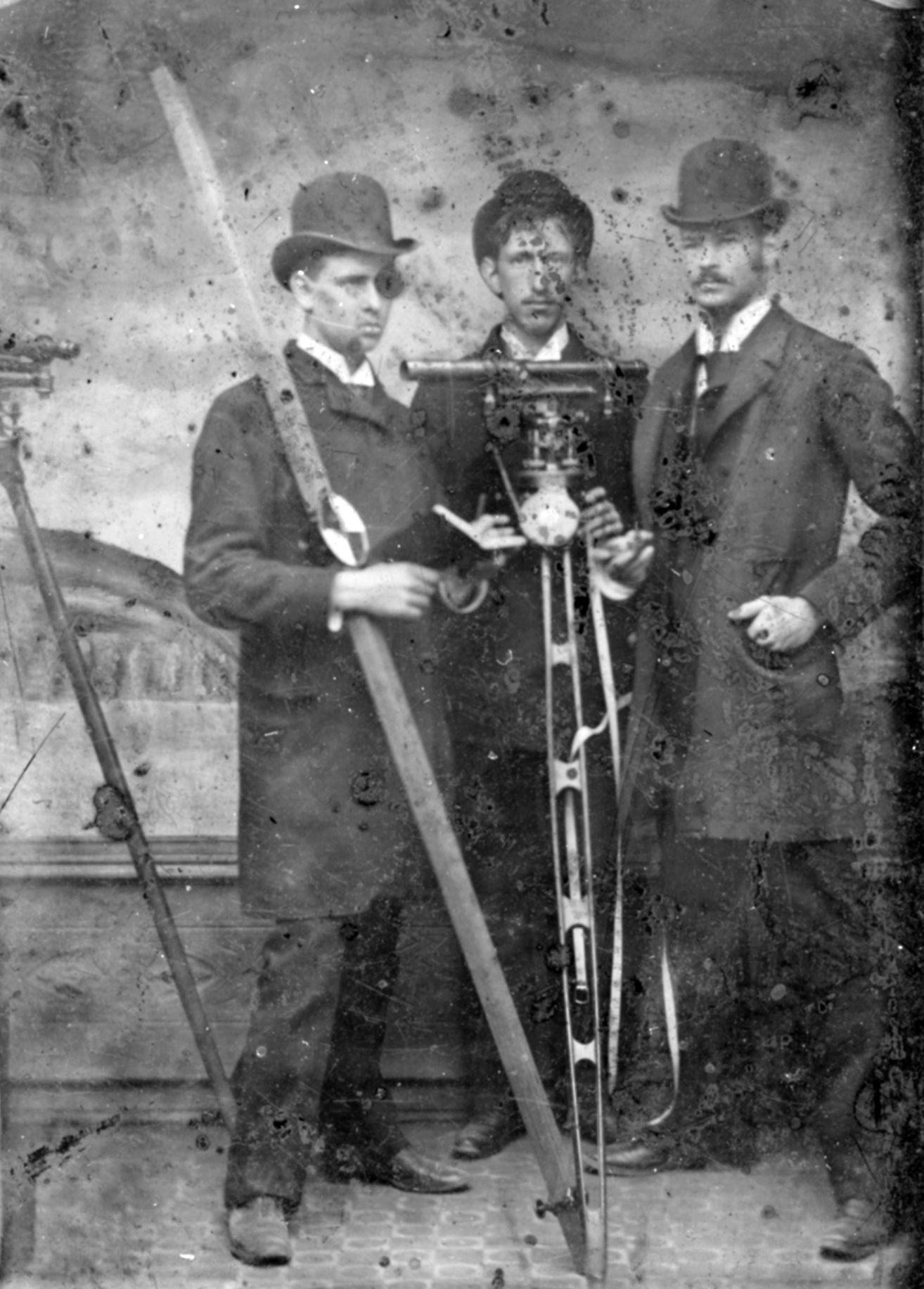 Black and white tintype photograph of three young men posed with surveying equipment.
