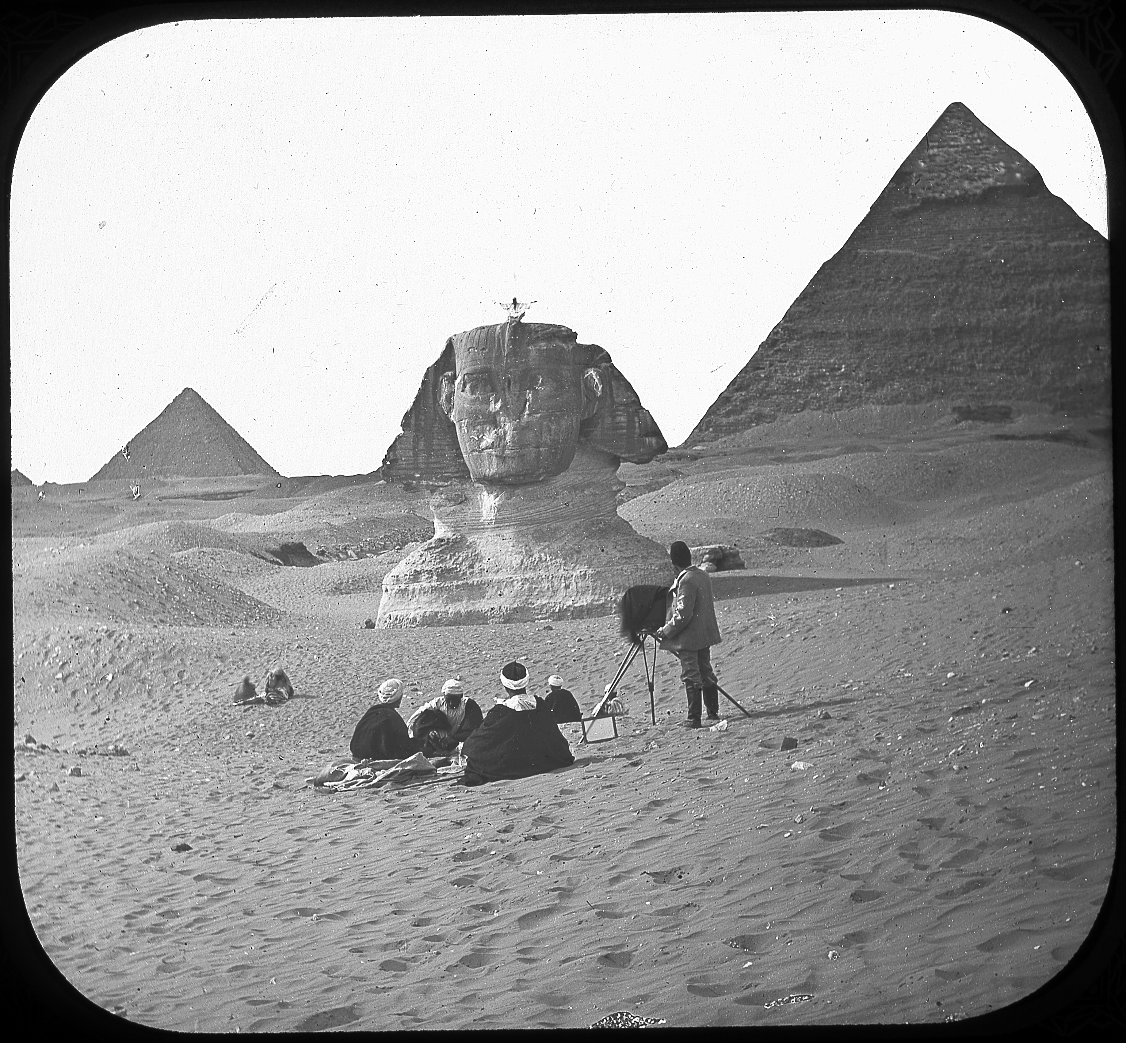 Black and white image of a photographer capturing a person sitting atop the at Sphinx, with the Pyramid of Chephren in the background