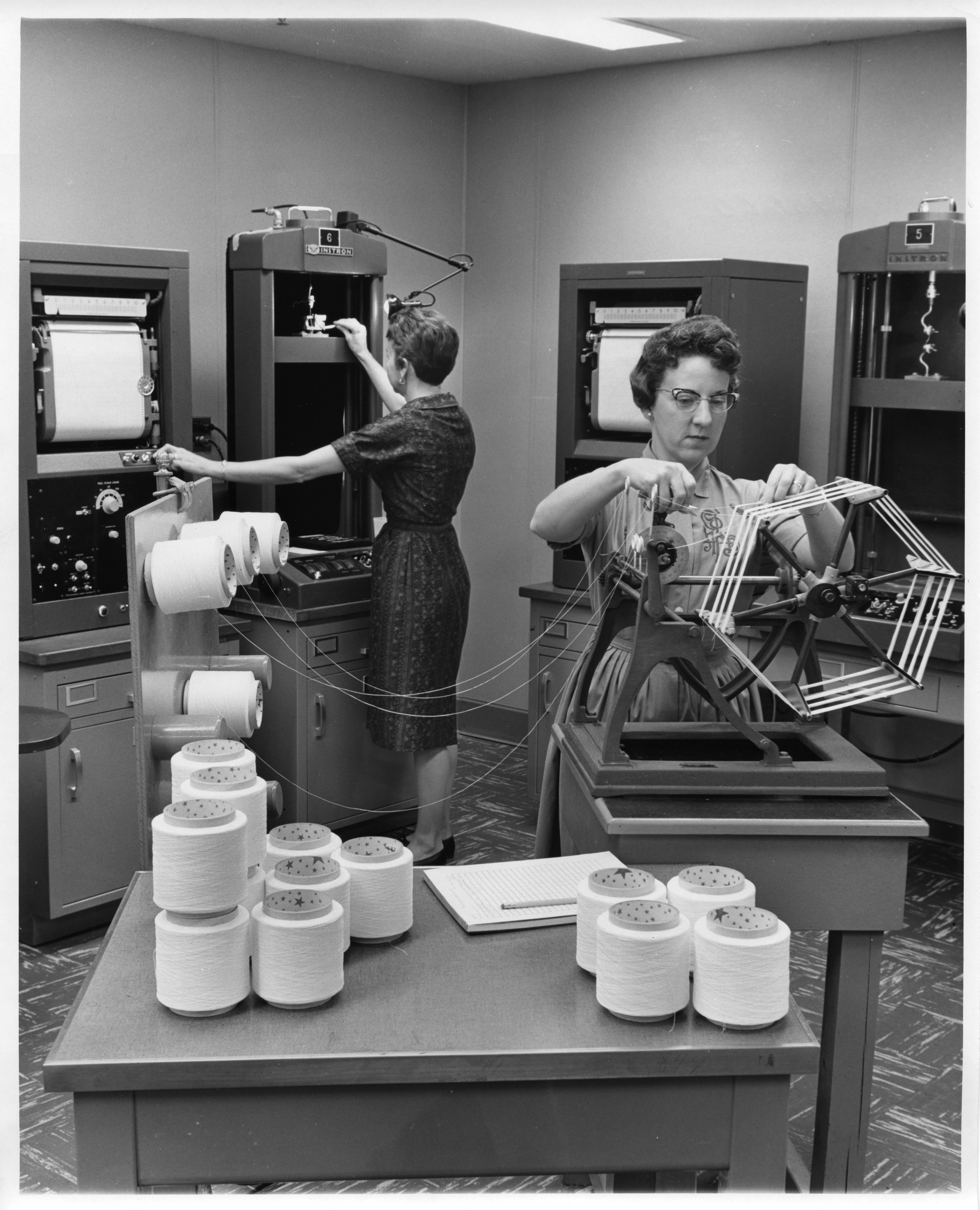 Black and white photograph of two women in a laboratory testing strands of Lycra fiber.