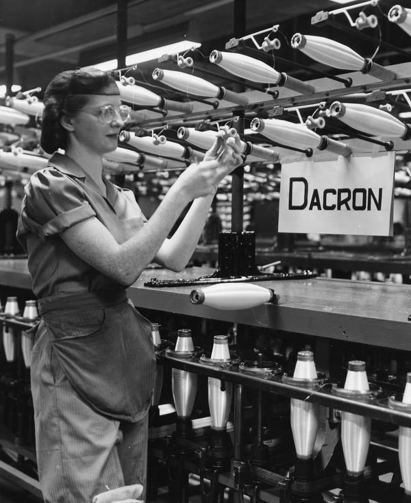 Black and white image of a woman working in front of an array of spools being filled with Dacron fiber. A sign nearby reads 'Dacron'