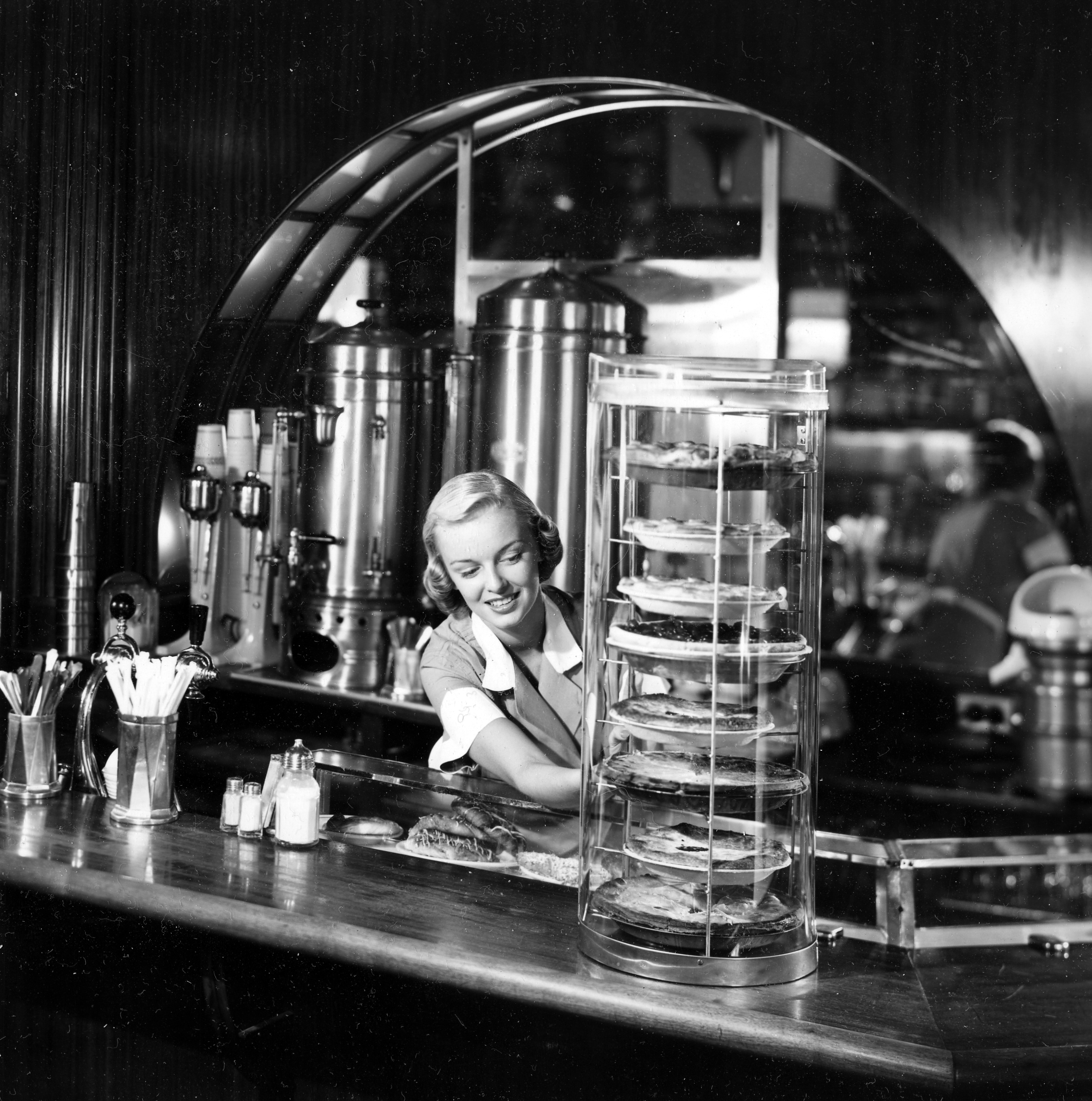 Black and white photograph of a woman at a diner adjusting a display case for pies.