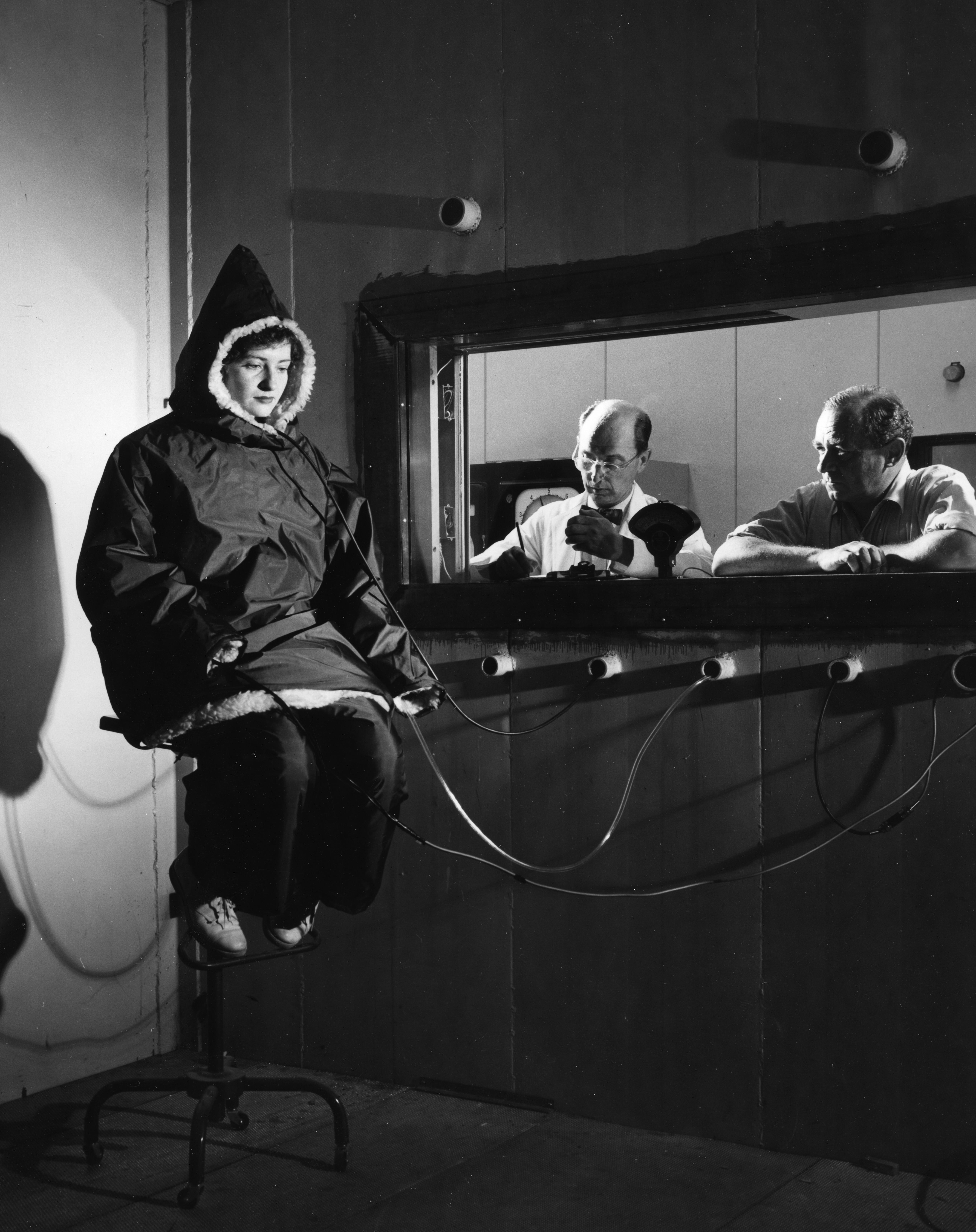 Black and white image of a woman in a large parka seated in a laboratory while two researchers observe behind a viewing window.