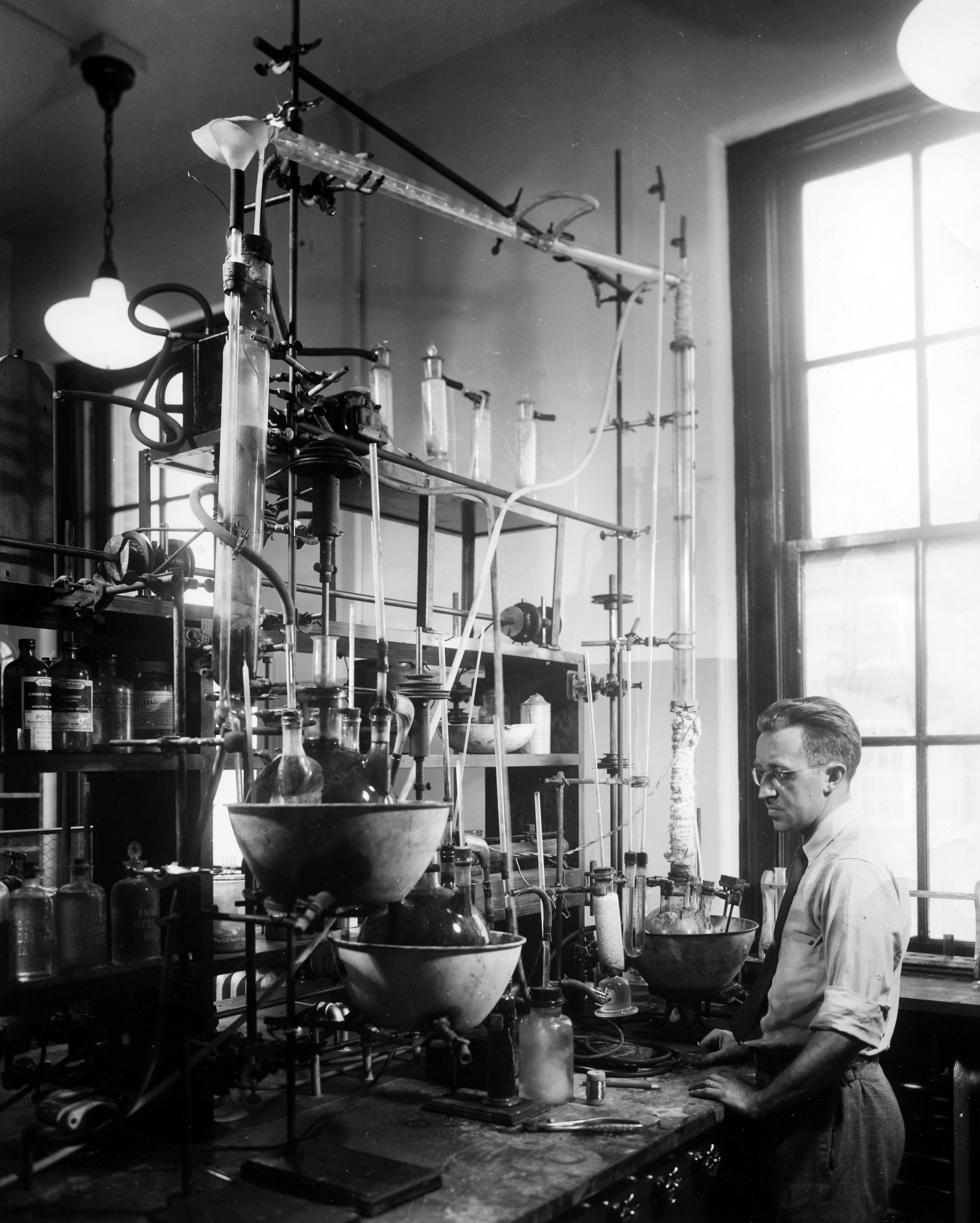 Black and white image of man standing in front of a large array of chemical laboratory apparatus.