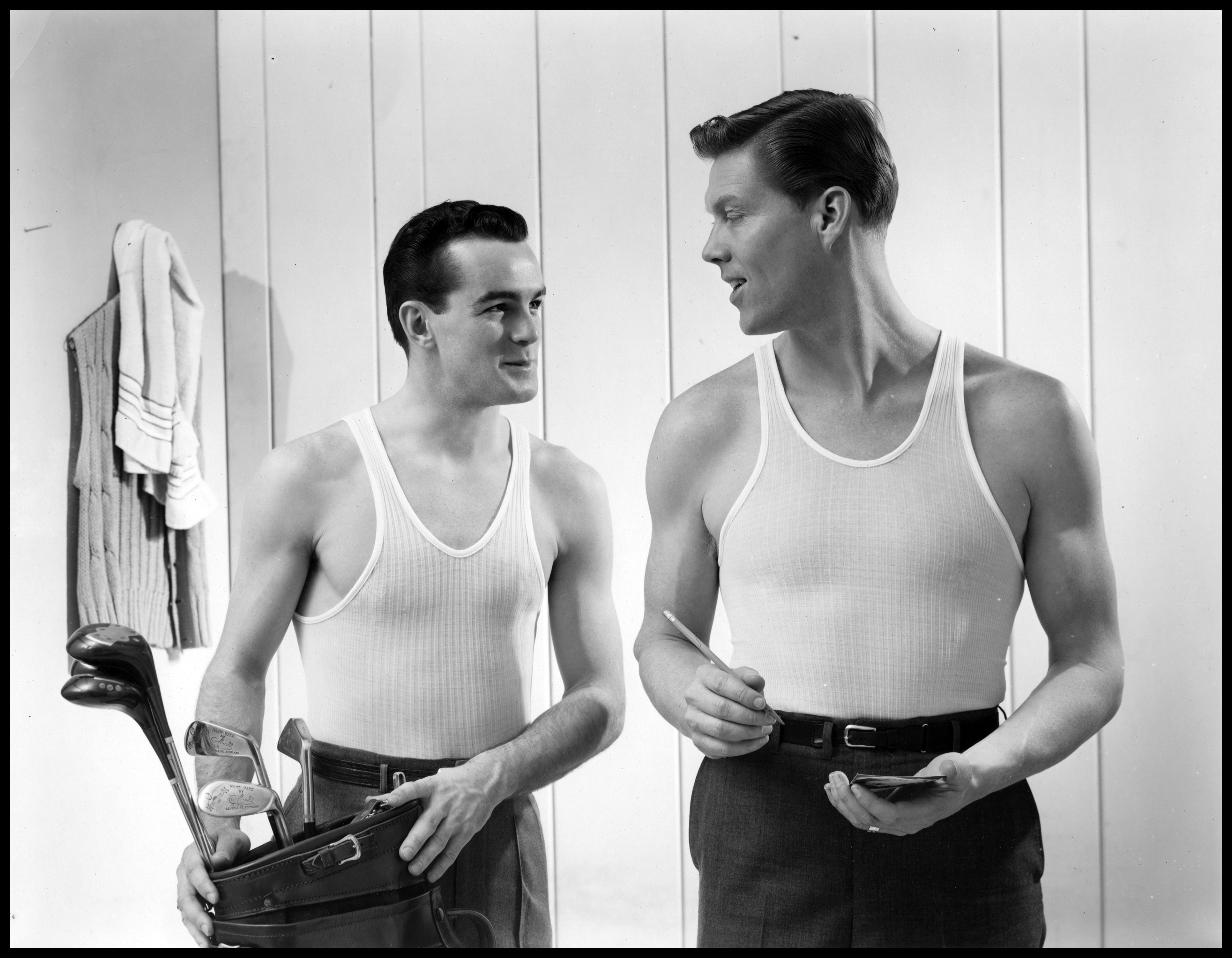 Black and white image of two men in undershirts with a golf bag.