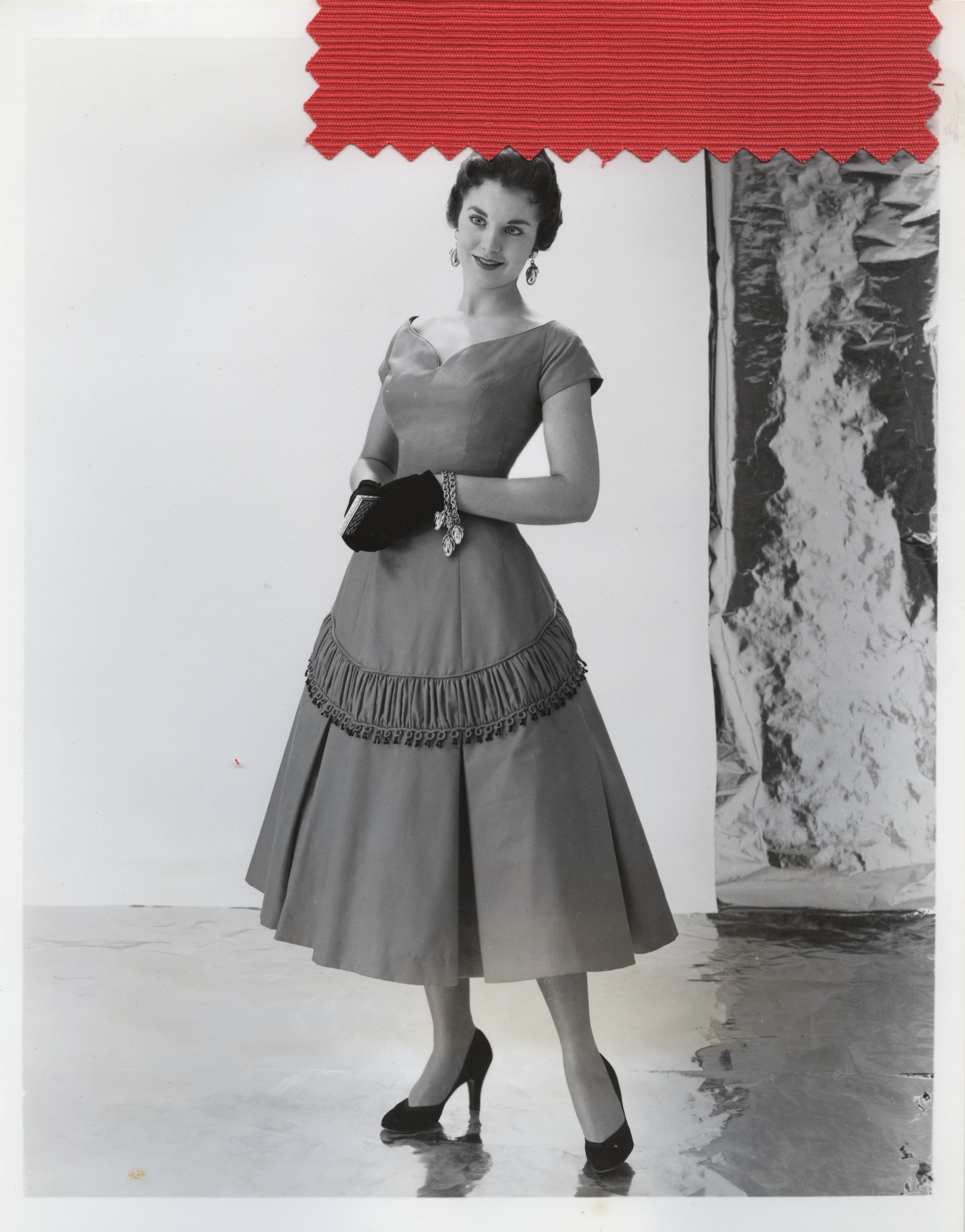 Black and white photograph of a woman in a calf-length cocktail dress, with a red fabric sample