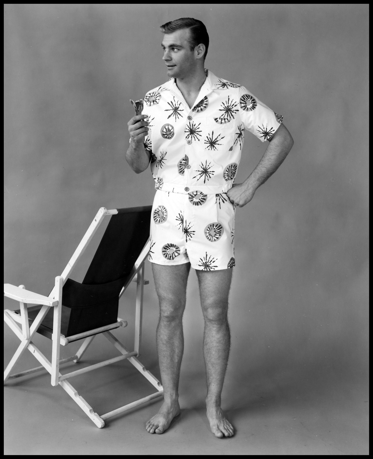Black and white image of a man in a "cabana set" outfit next to a beach chair on a photography set.