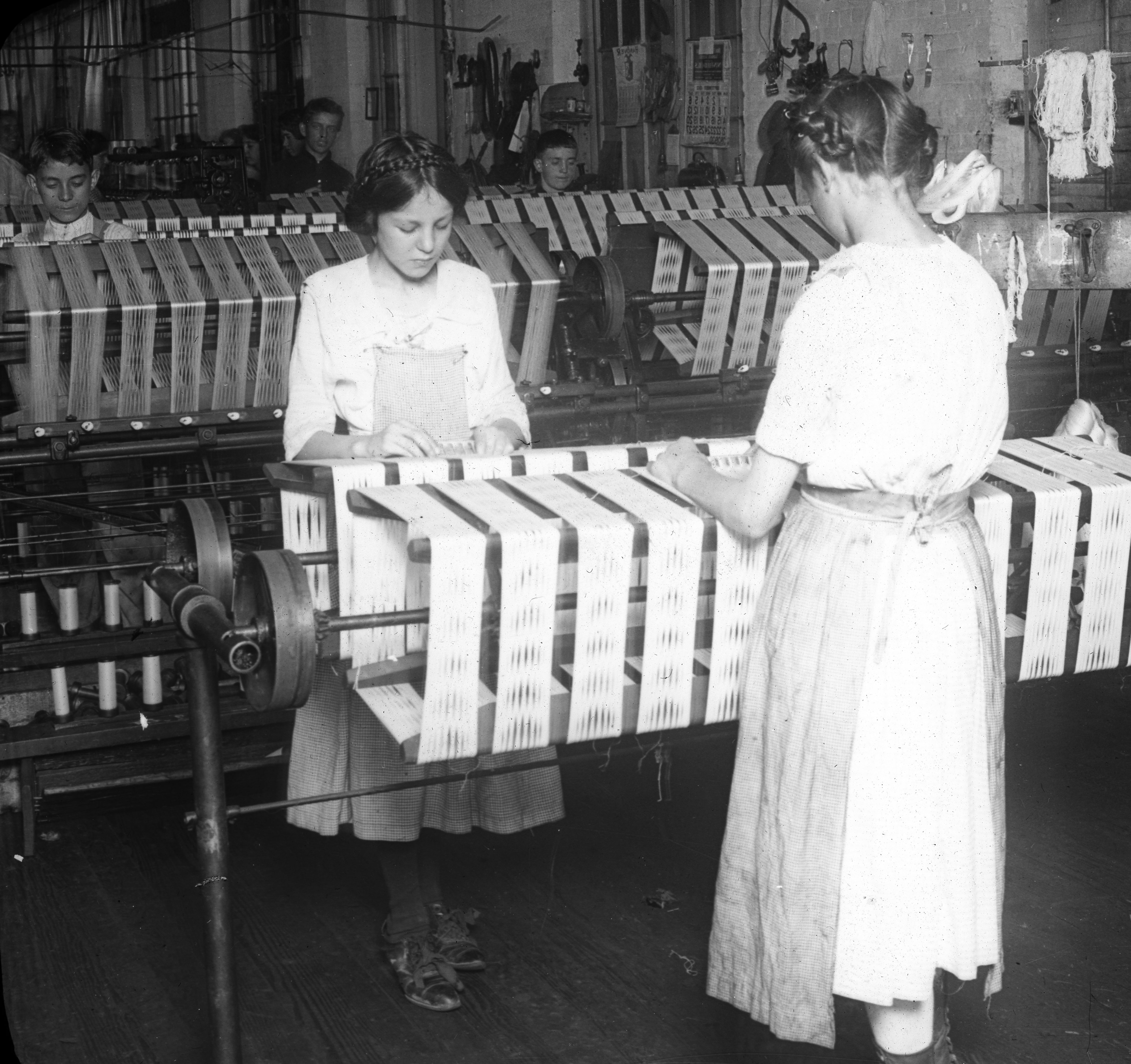 Black and white image of two young women working at a stand of silk manufacturing machinery.