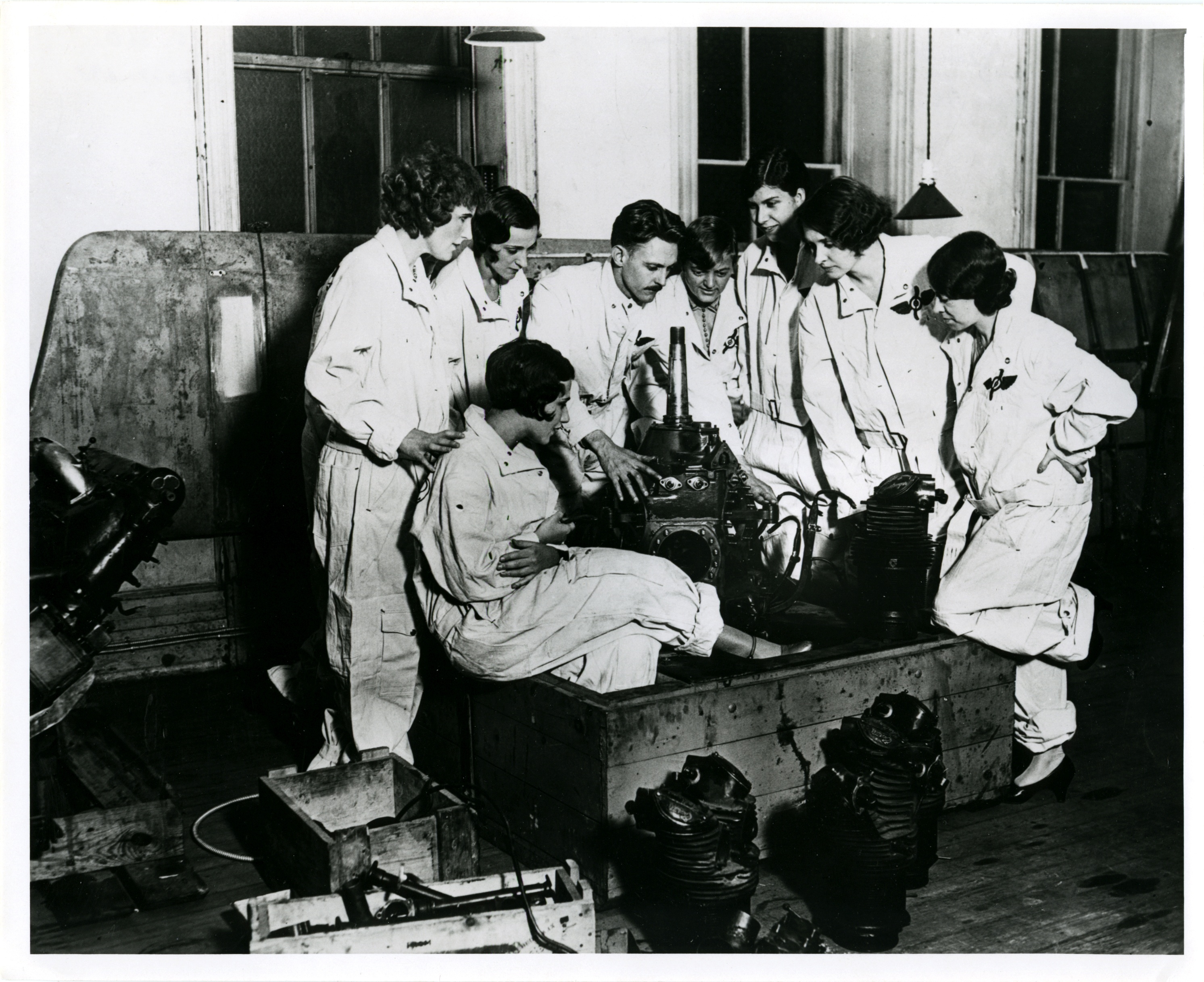 Group of women in flights suits gathered around men demonstrating the parts of an aircraft motor.