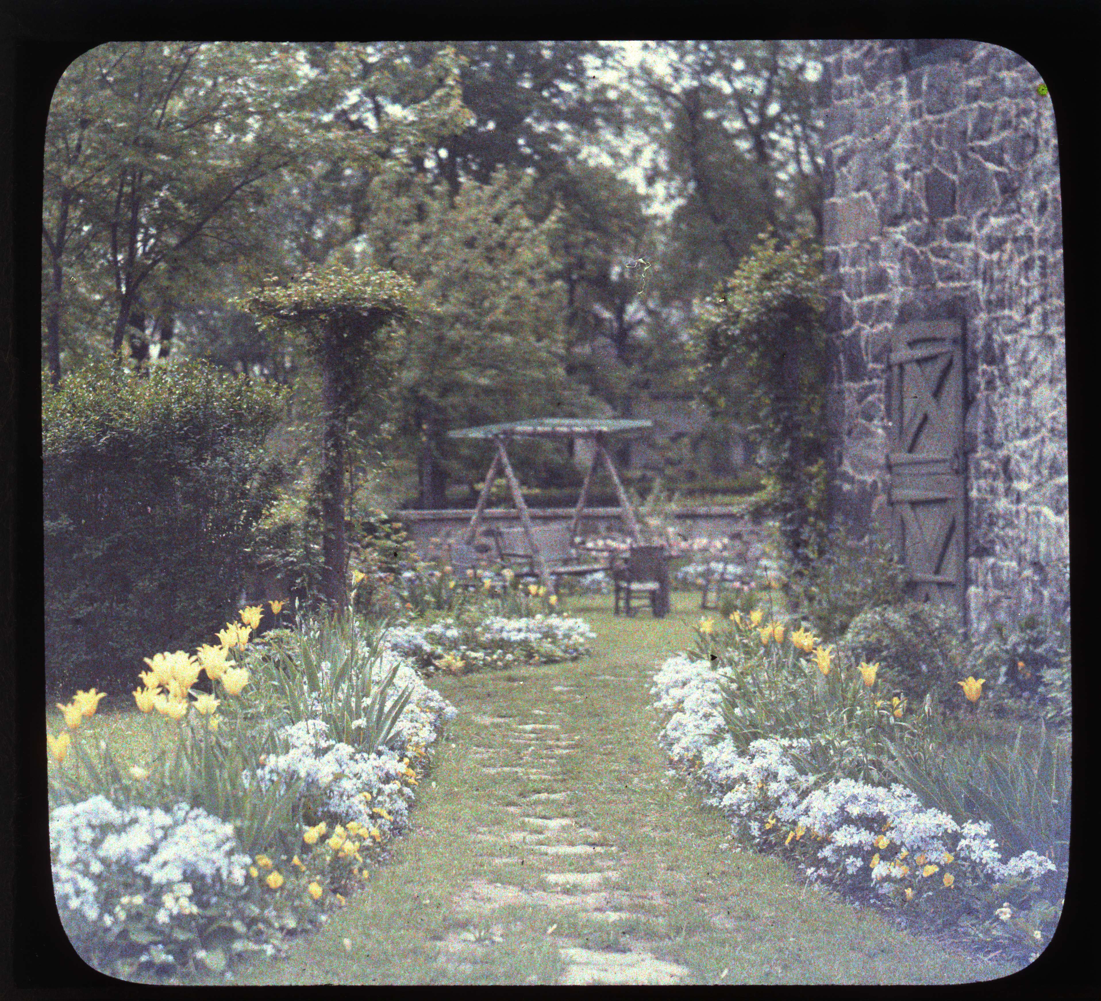 Autochrome image of a garden in bloom with a stone building and garden swing