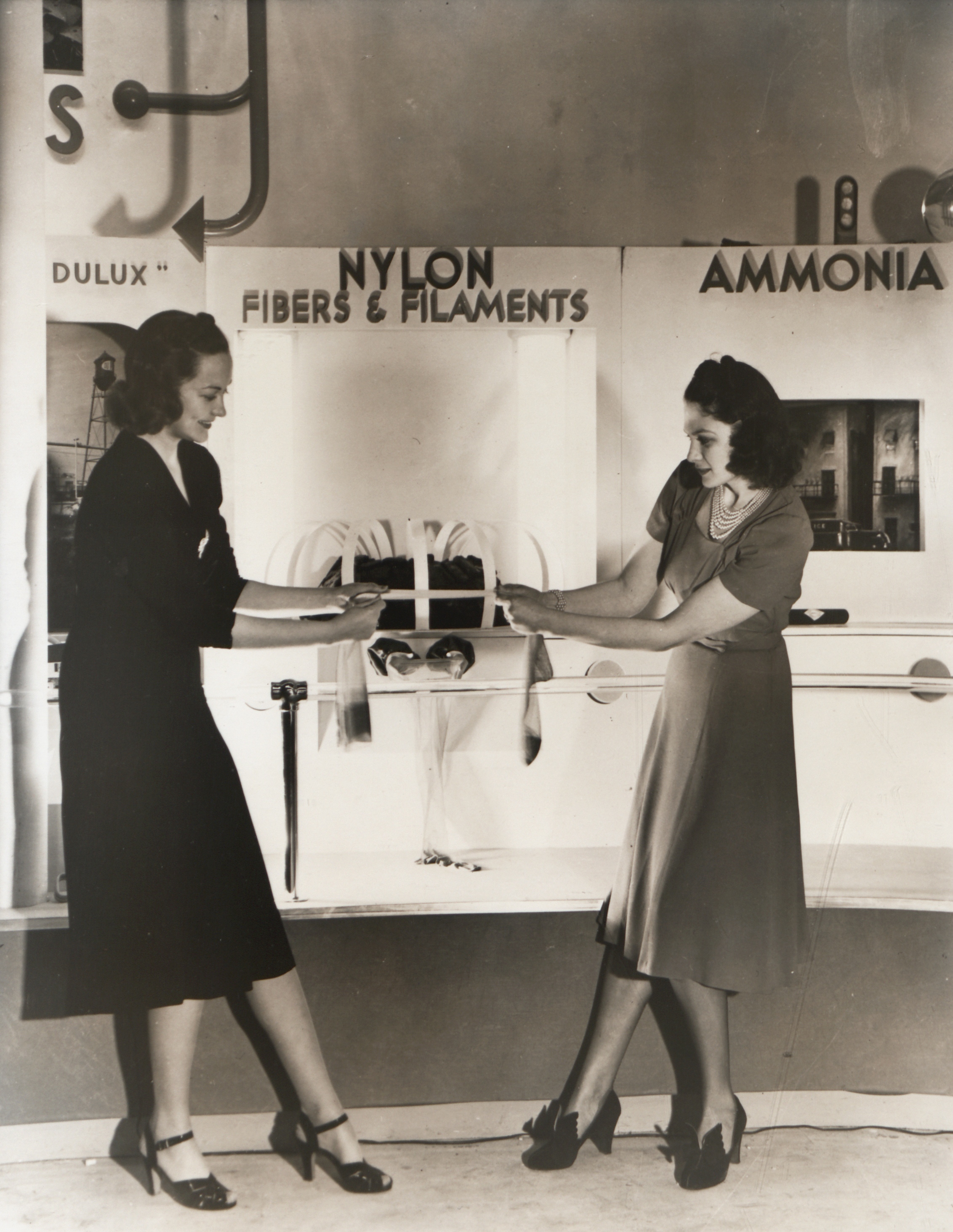 Black and white image of two women at an exhibition booth pulling on either end of a leg of nylon stocking.