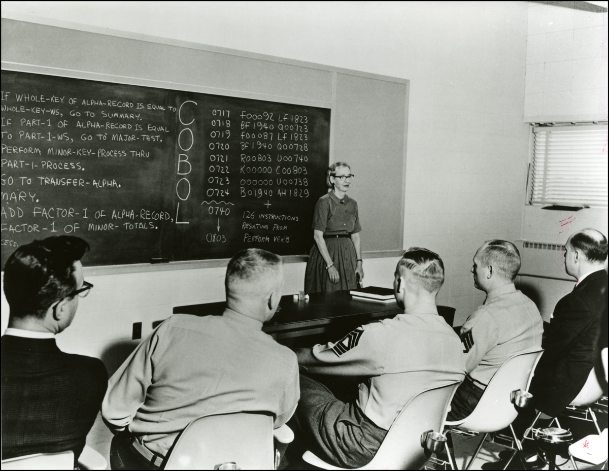 Black and white image of Grace Hopper presenting on COBOL in front of blackboard