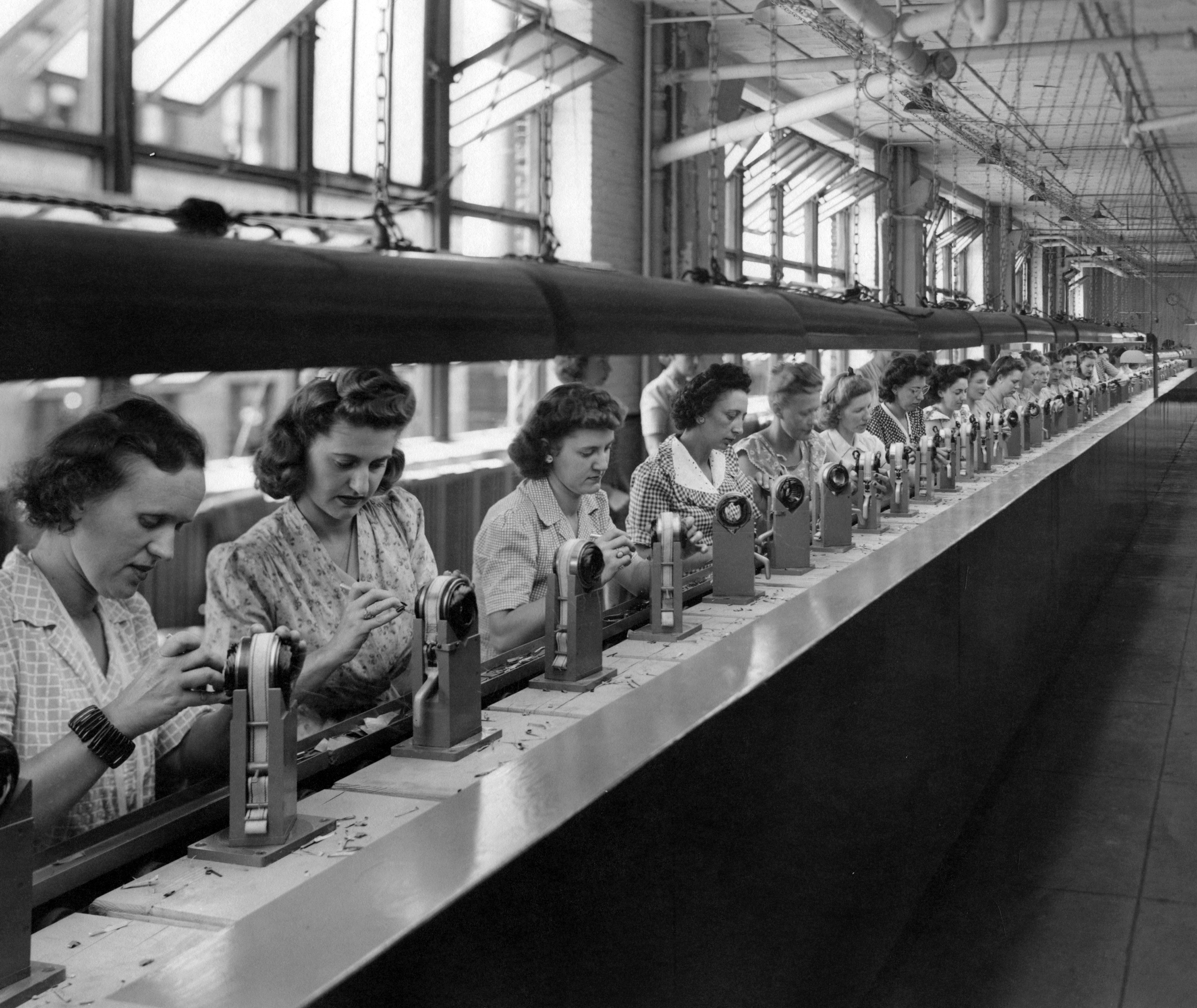 Photograph of a long line of women in a factory, each working on a machine part in front of them on a conveyor belt.