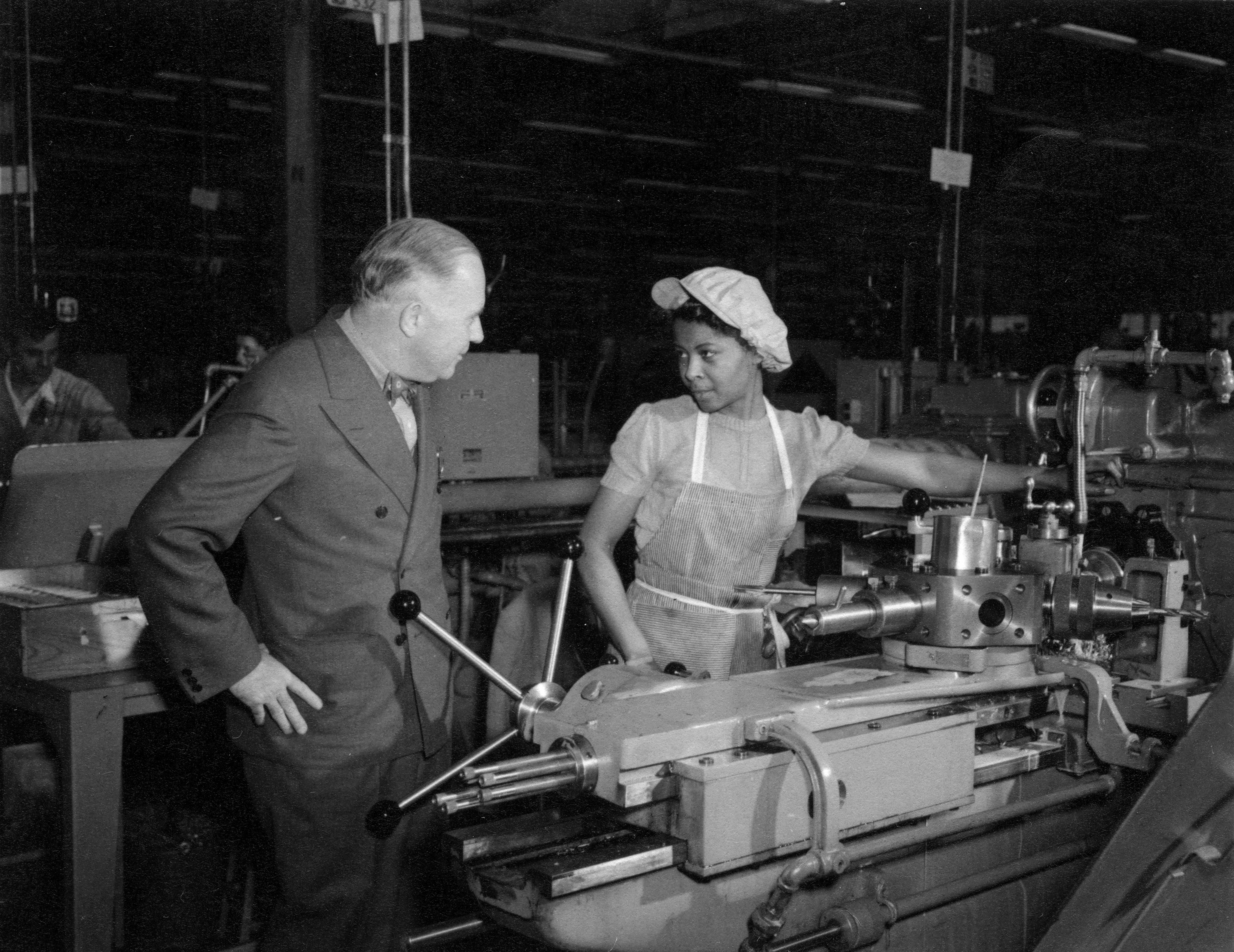 Black and white photograph of a man speaking to a woman operating a lathe.