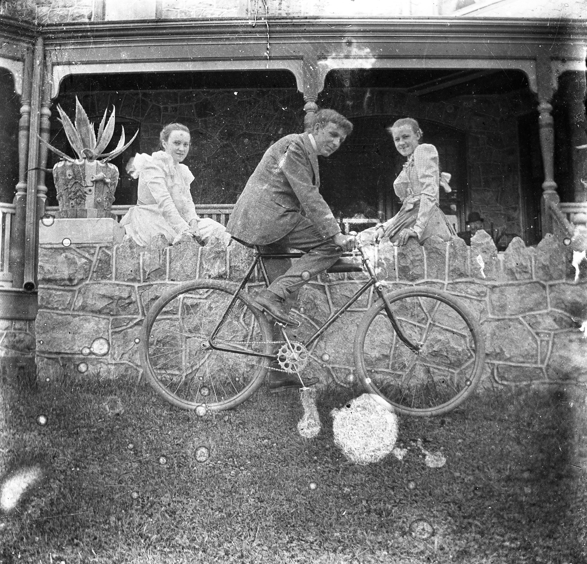 Black and white negative photograph of a man on a bike, flanked by two women sitting on a wall.
