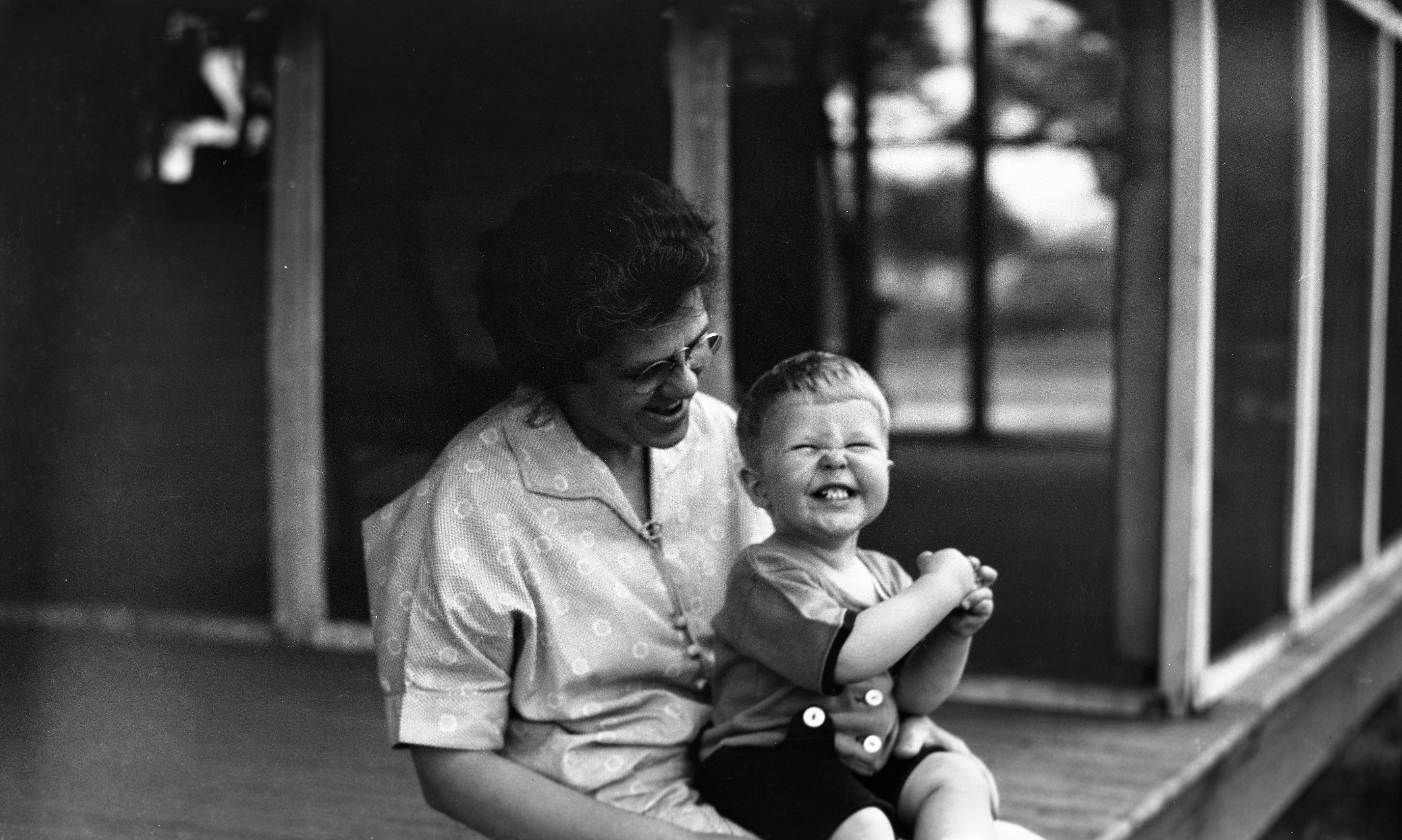 Black and white image of a smiling toddler seated on a woman's lap on a porch