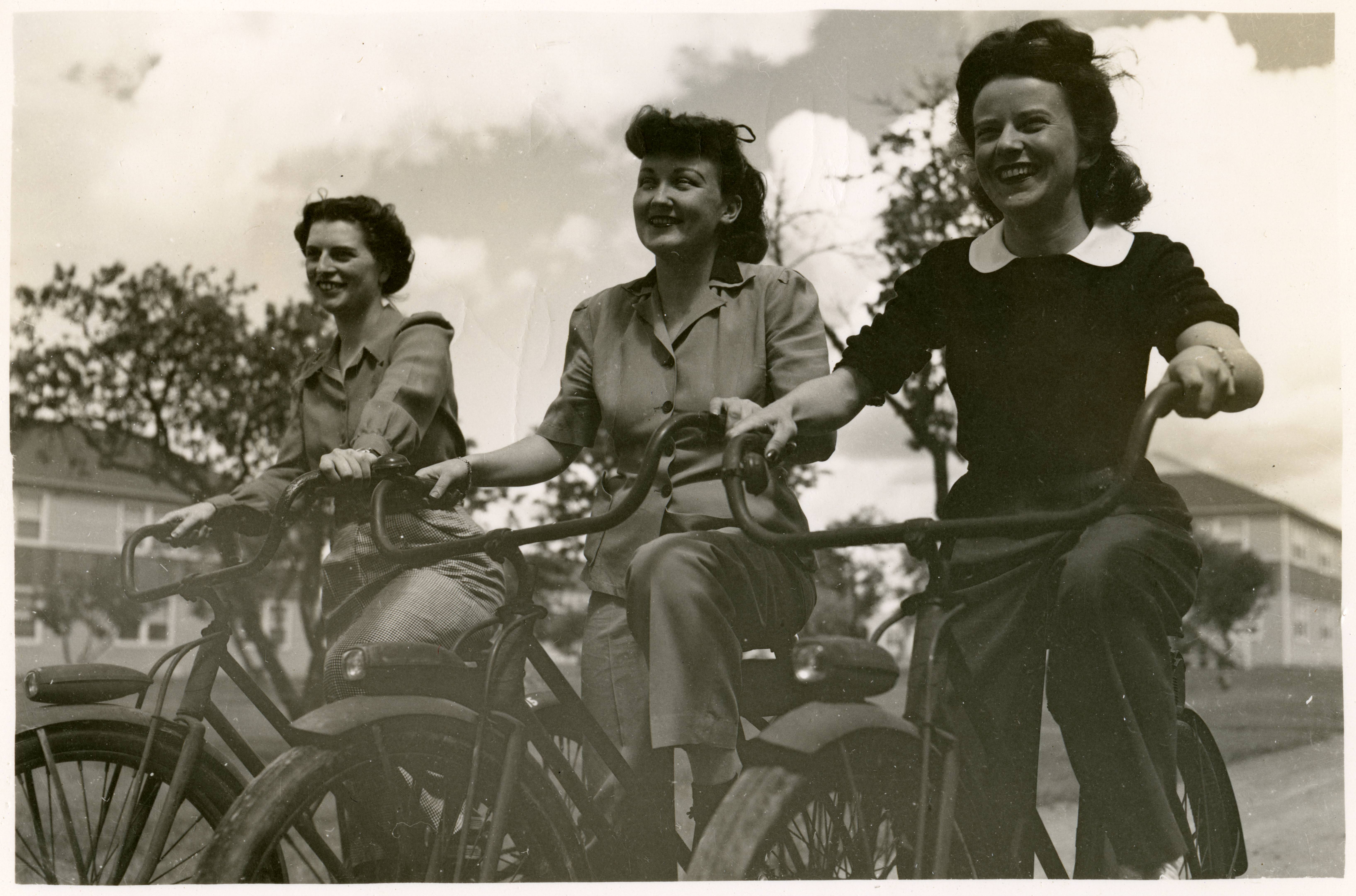 Black and white photograph of three women in work clothes riding bicycles.