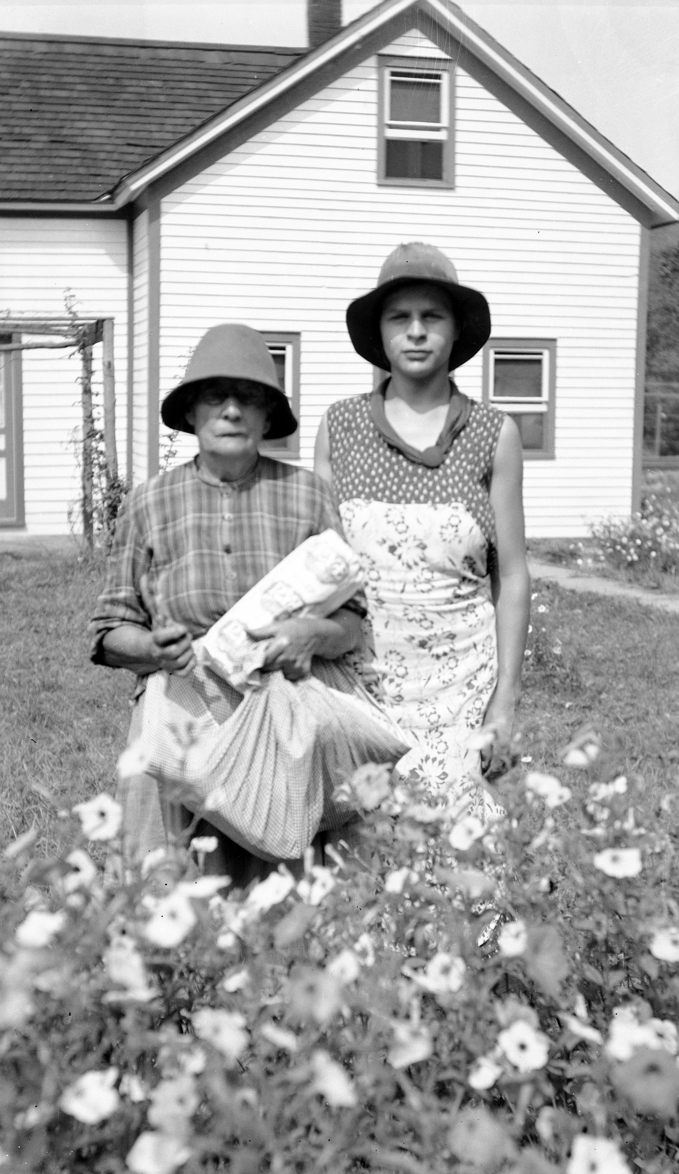 Black and white photo. Two people posed behind a waist-high flowering plant. Home and yard in the background.
