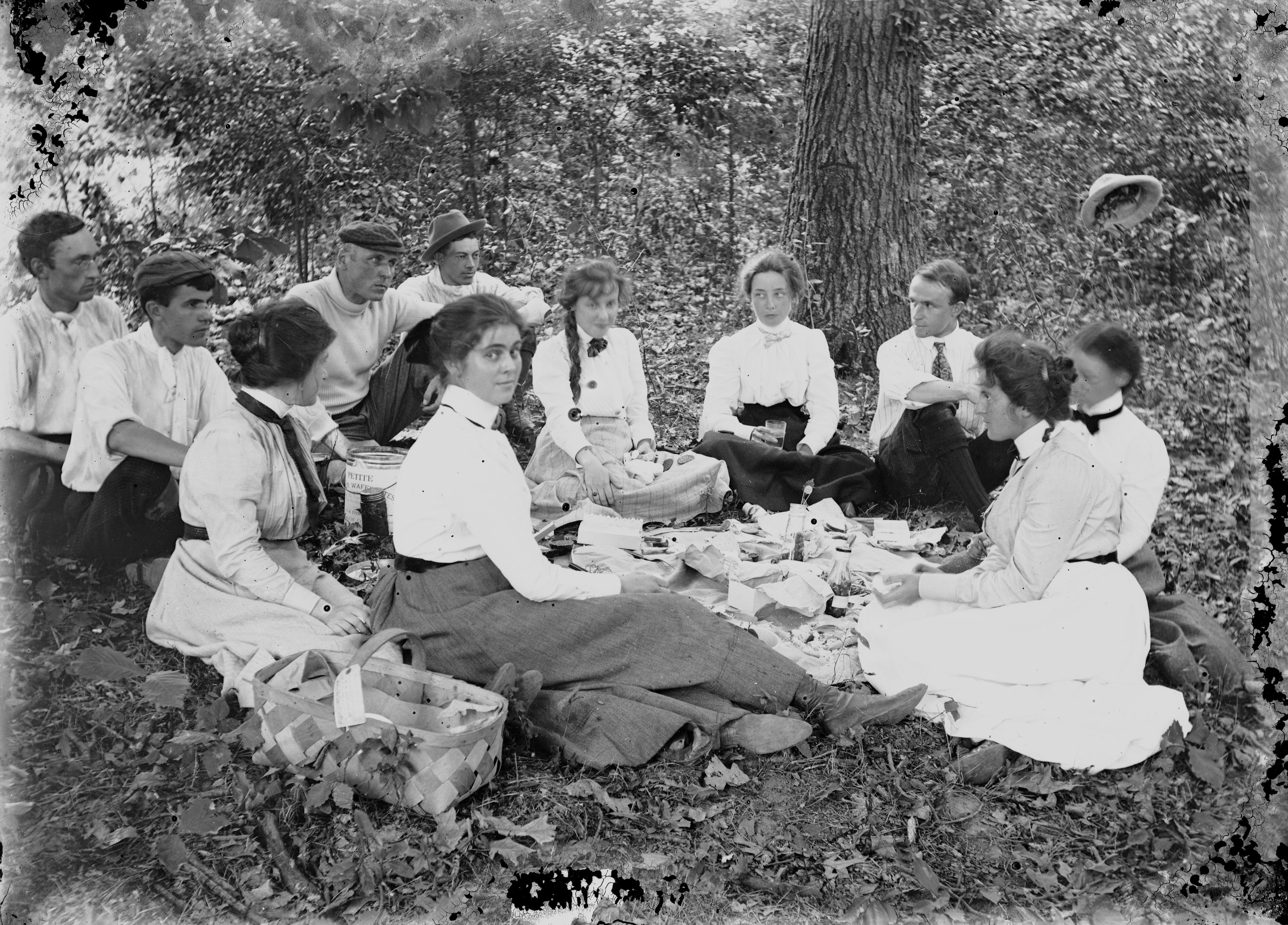 Black and white glass negative showing a group of young men and women having a picnic in the woods. An older man, artist Howard Pyle, is also present.