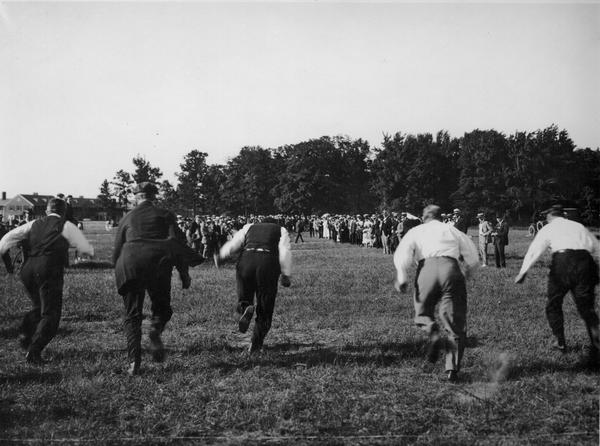 Black and white photo of men running, captured from behind, crowd in distance.