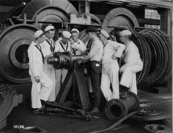 Black and white image of cadets dressed in nautical uniforms examining large machinery in a factory..