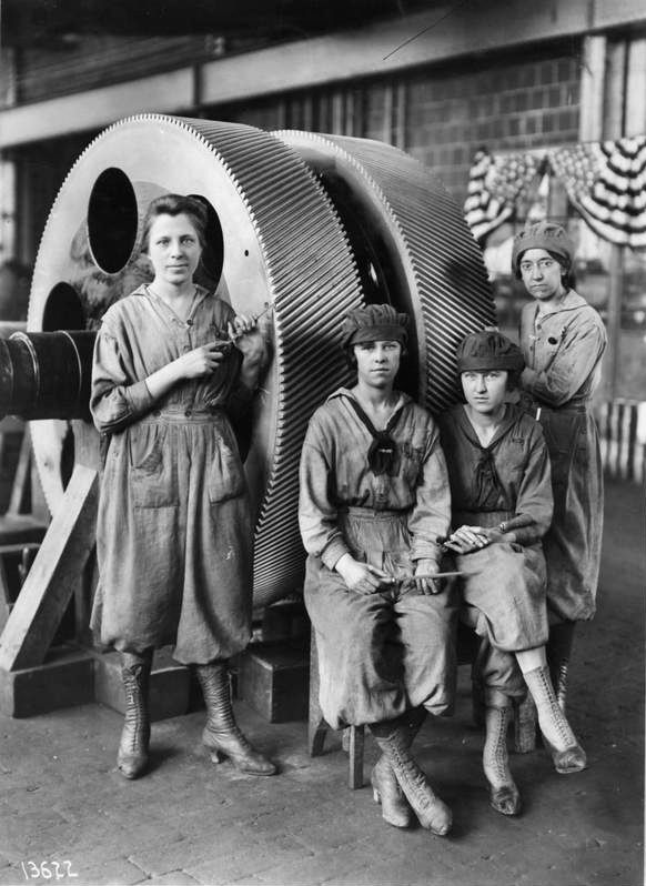 Black and white photograph of four women in work clothes, standing in front of large industrial gears.