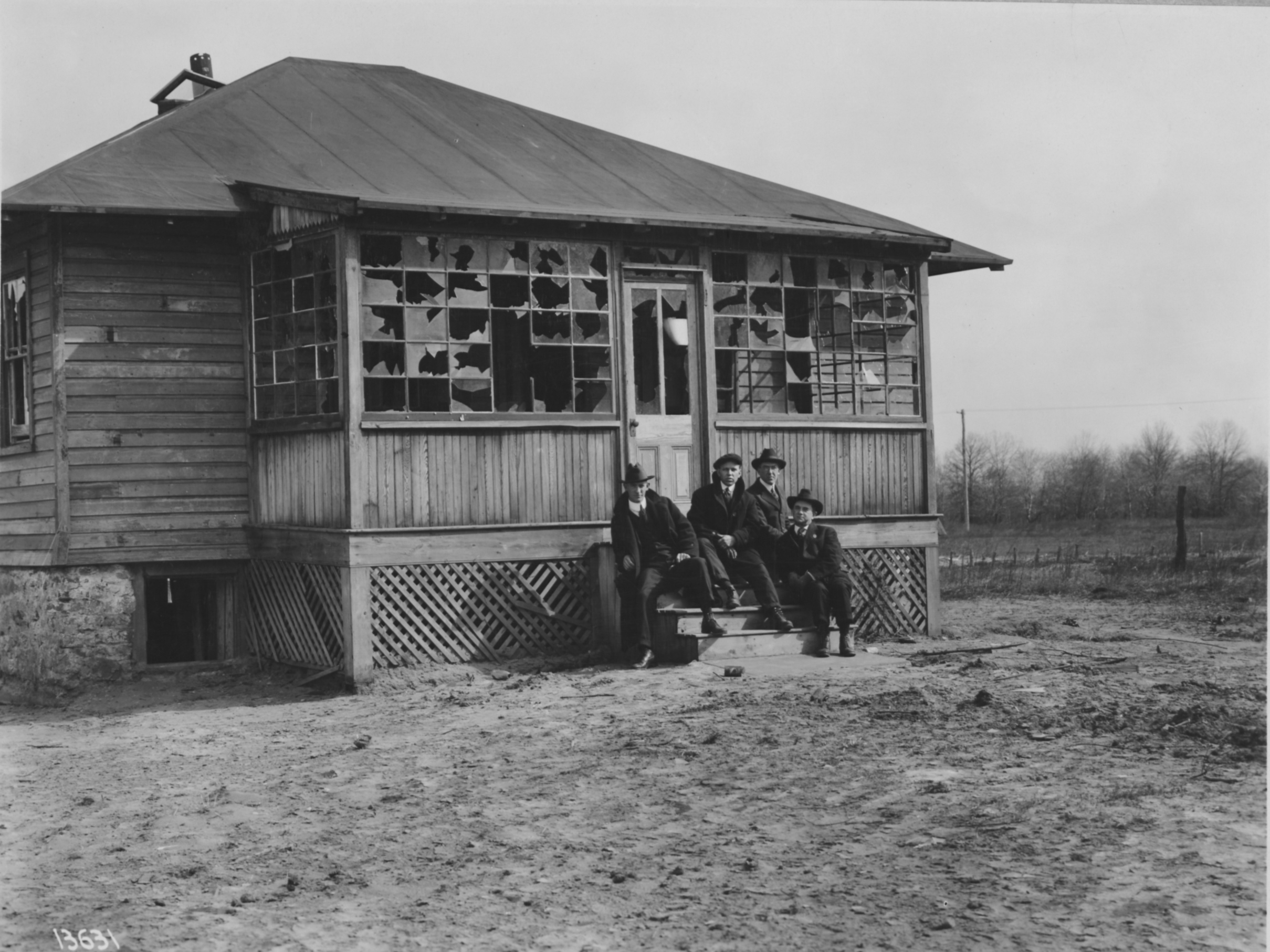 Black and white image of four men in hats and suits seated on the steps of a small building in poor repair.
