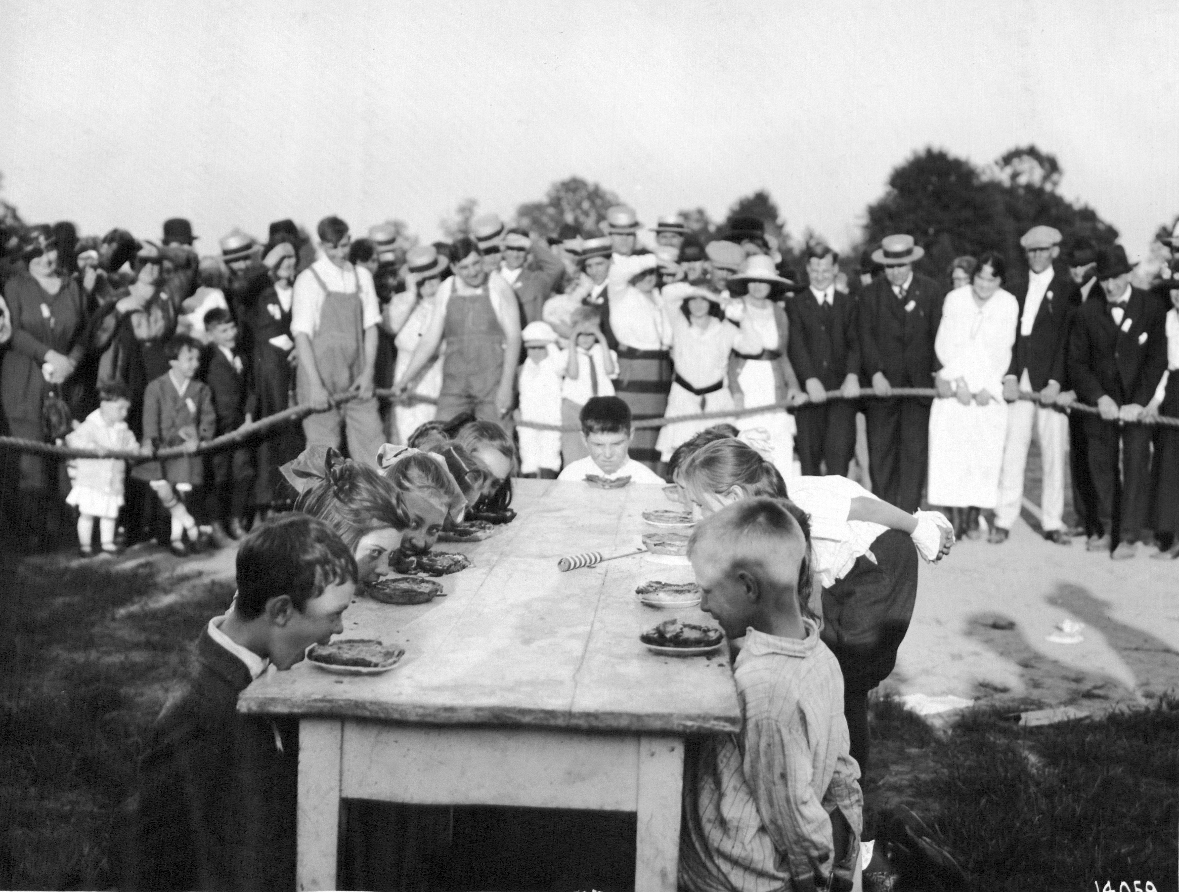 Black and white image of children lined up at a table, in front of an audience, for a pie eating contest.
