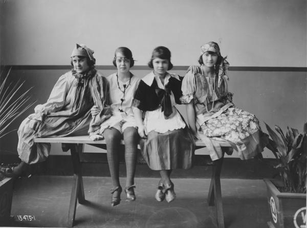 Black and white photograph of four young women in costumes, seated on a wooden table.