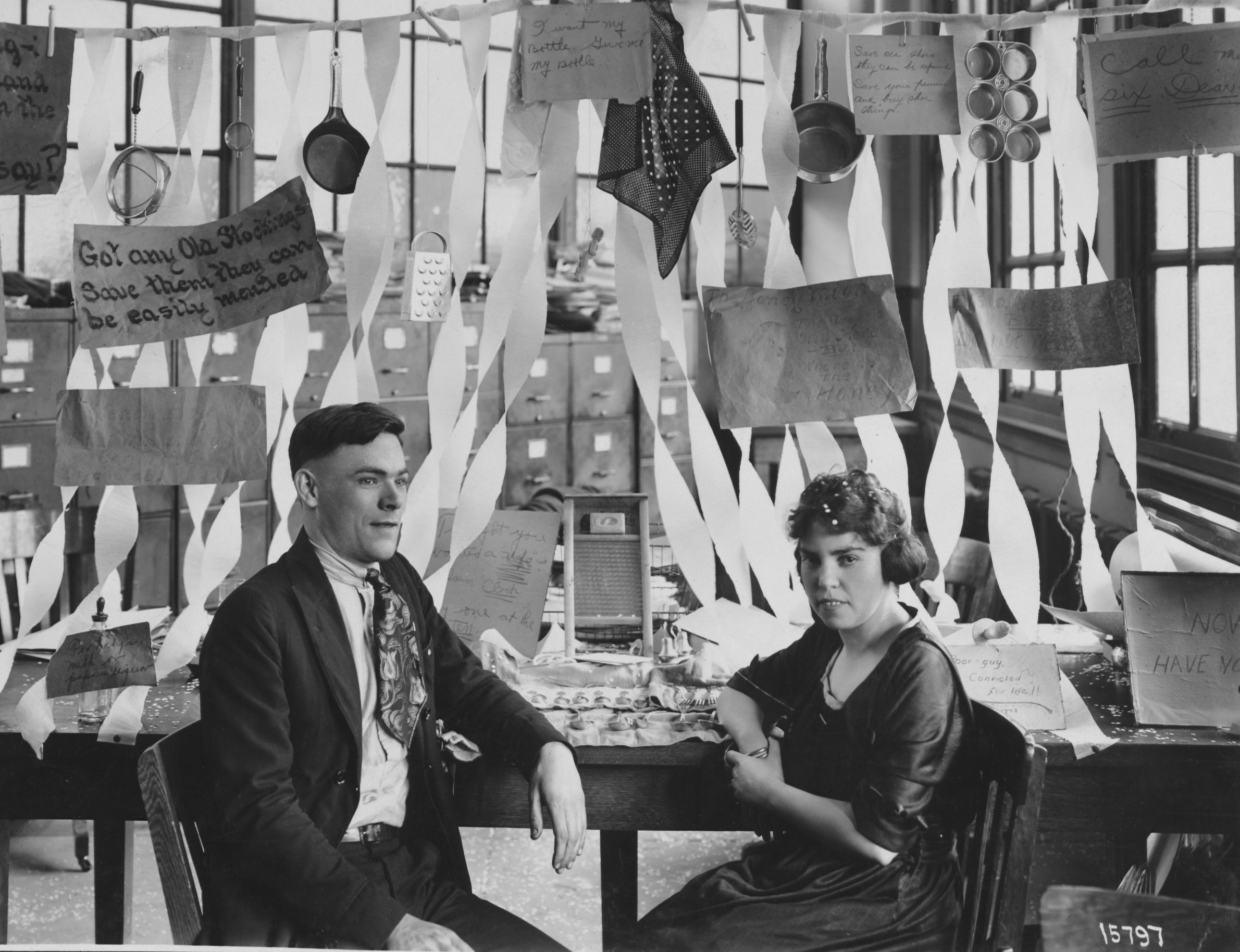 Black and white photograph of a man and woman seated in front of a desk decorated with a wedding theme.