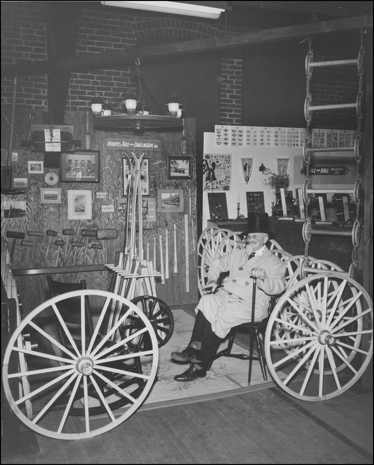 Black and white image of a man in a coat and top hat, seated in a booth exhibiting the works of the Hoopes Brothers & Darlington company. 