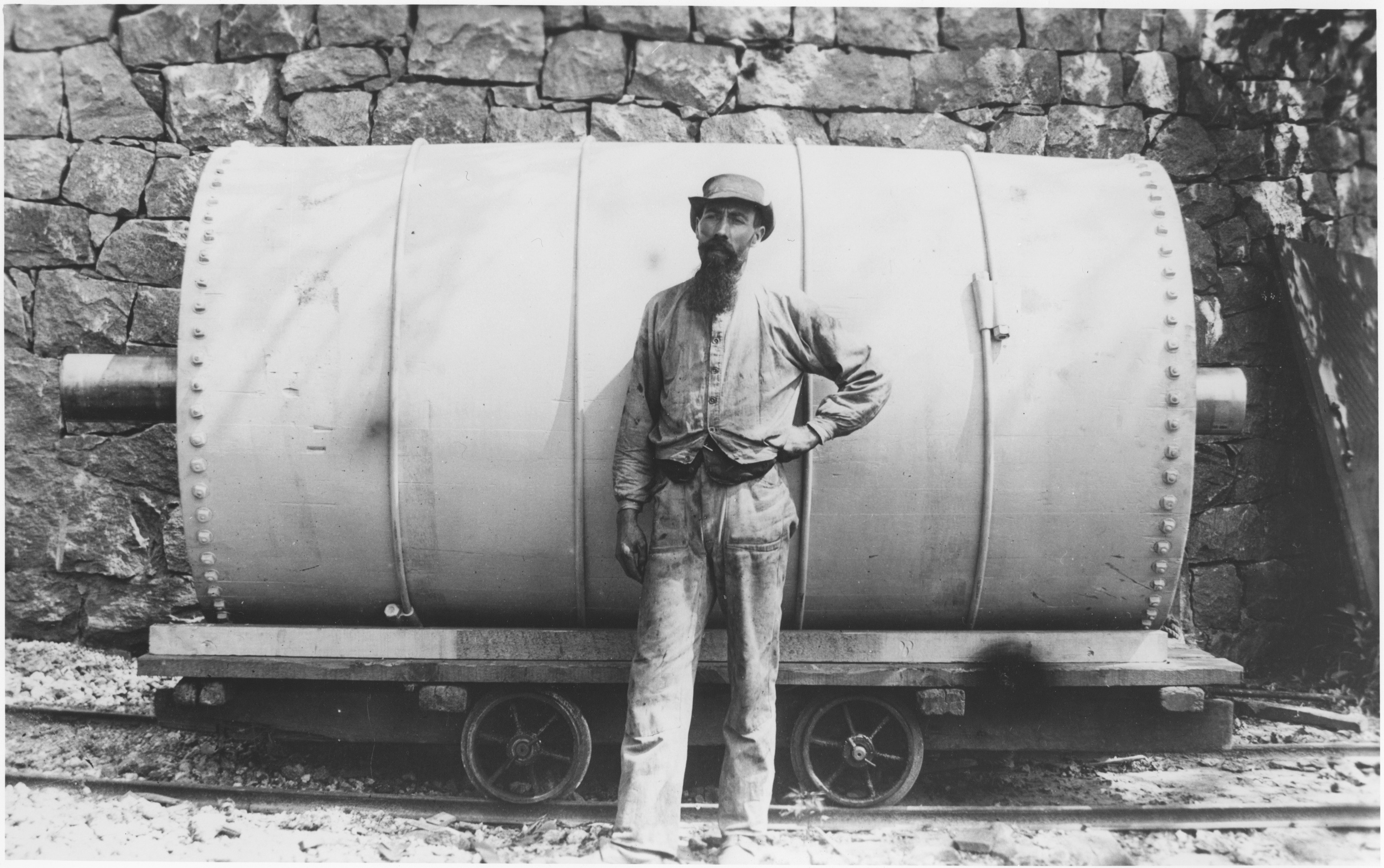 Black and white image of a bearded person standing in front of an industrial glazing barrel.