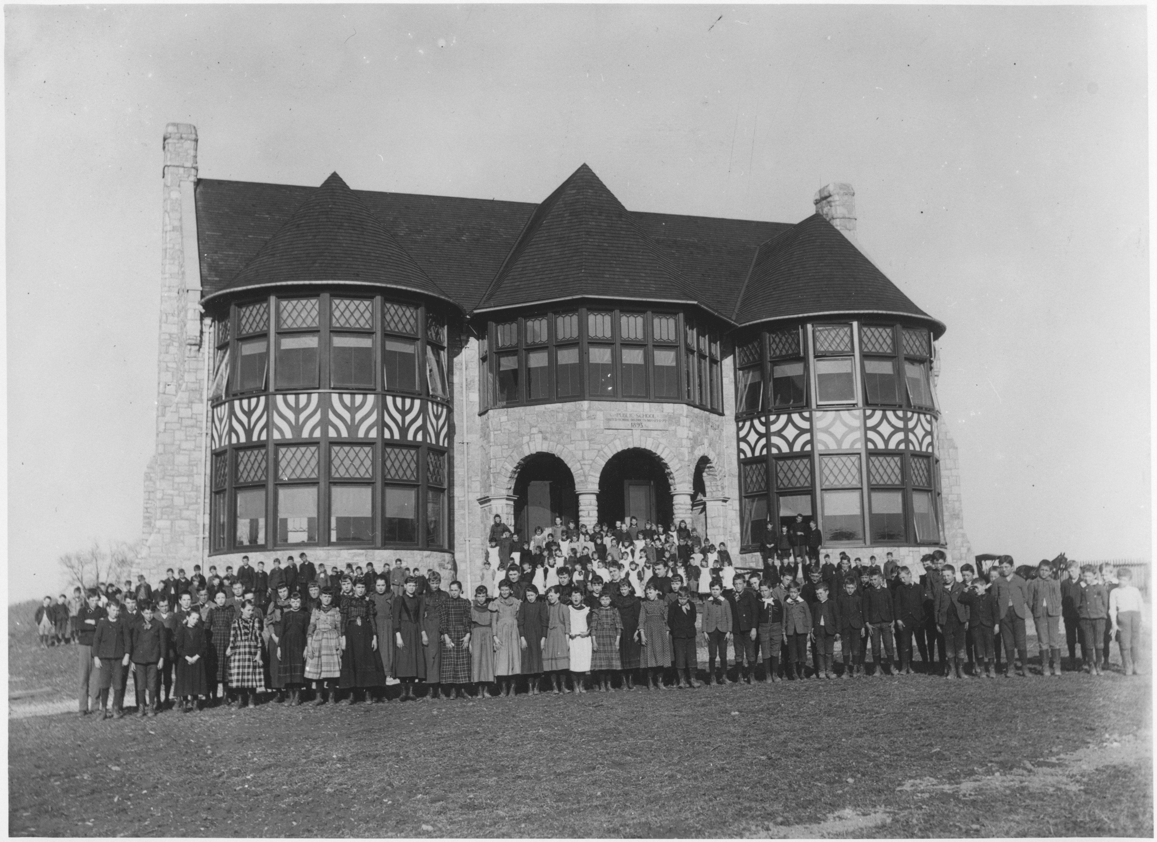 Black and white group photograph of many children posed outside of a large school building