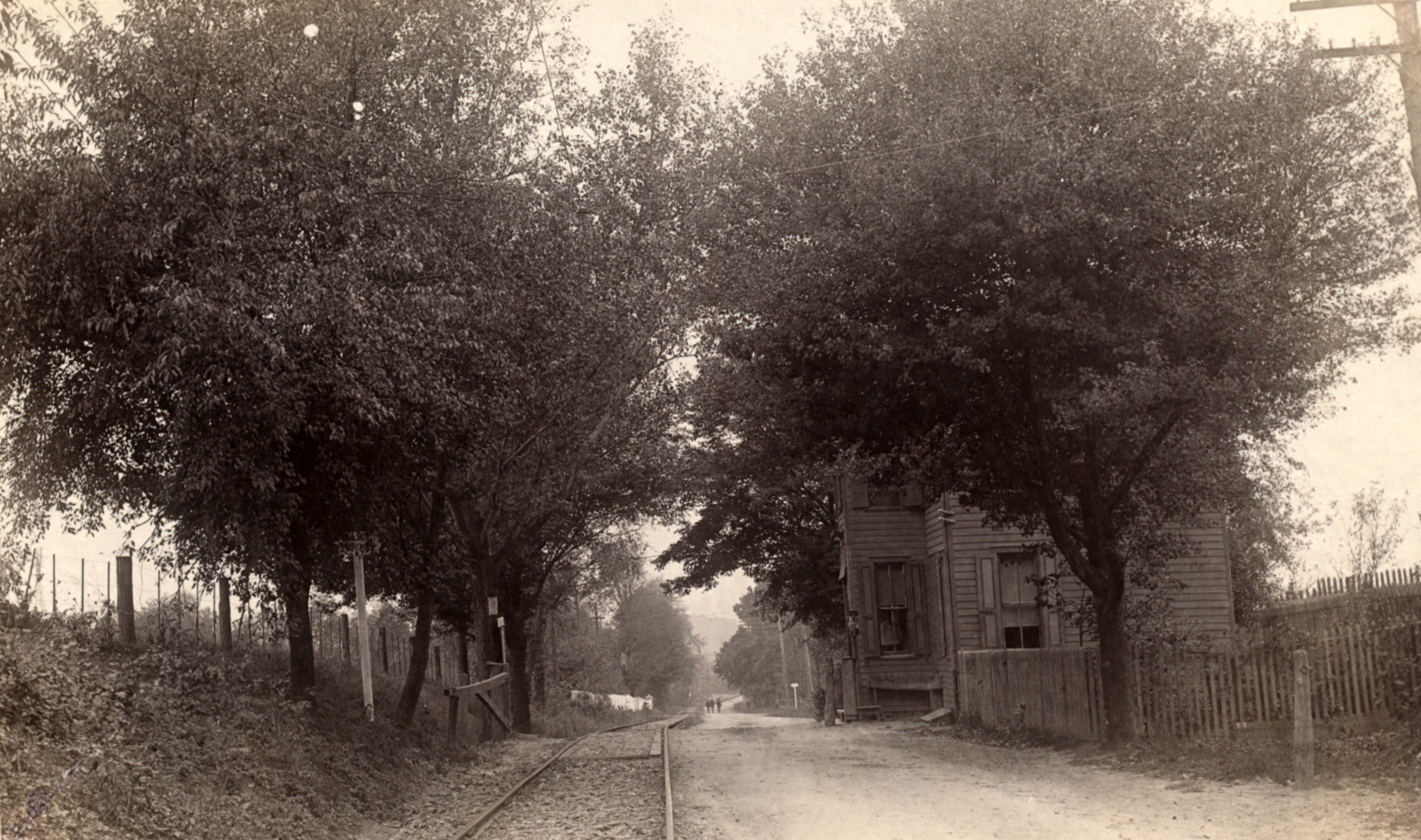 View of a turnpike and trolley line, running alongside a tollhouse and tollgate, in a rural setting.