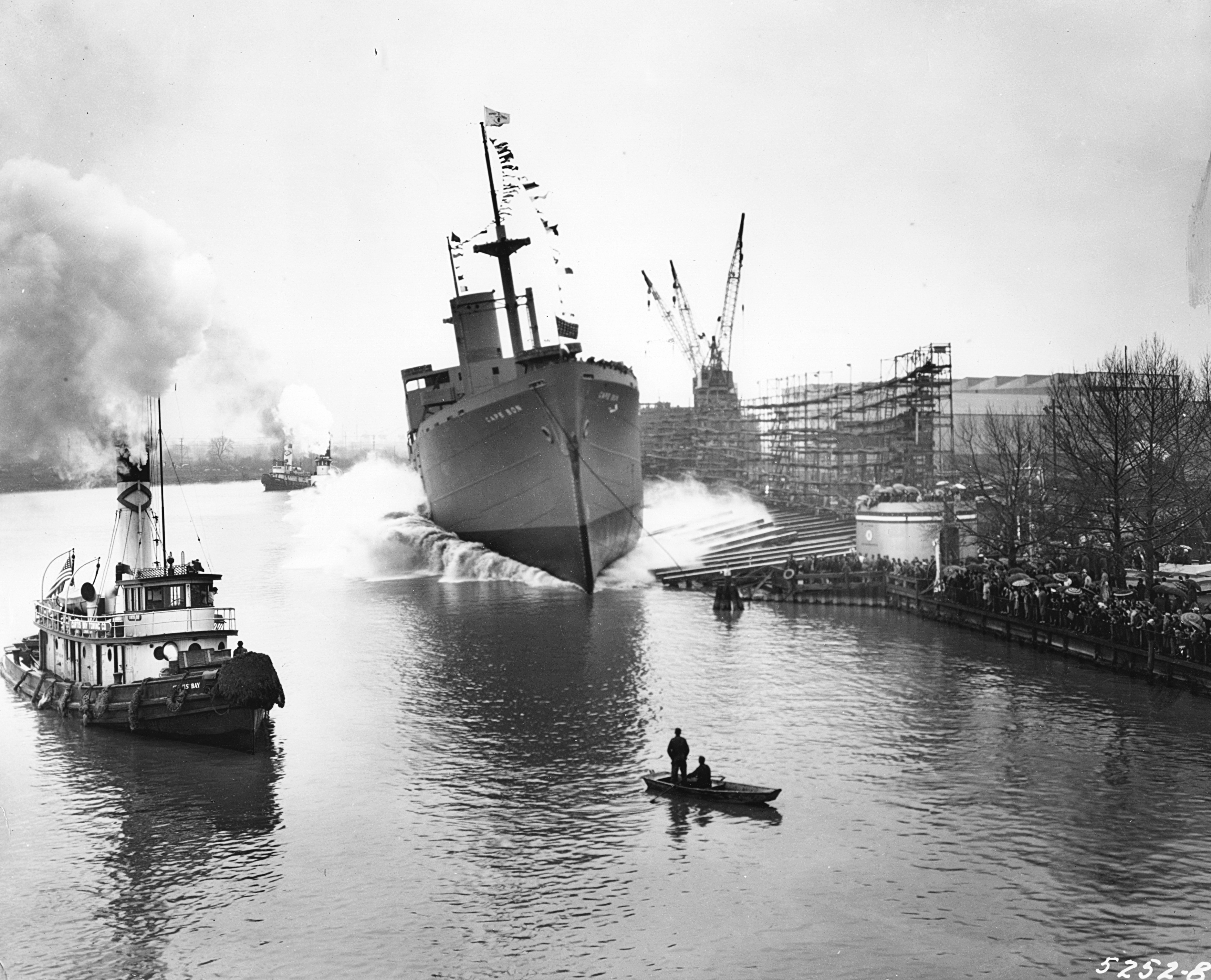 Black and white image of a large freighter ship being dropped into a busy river with many ships.