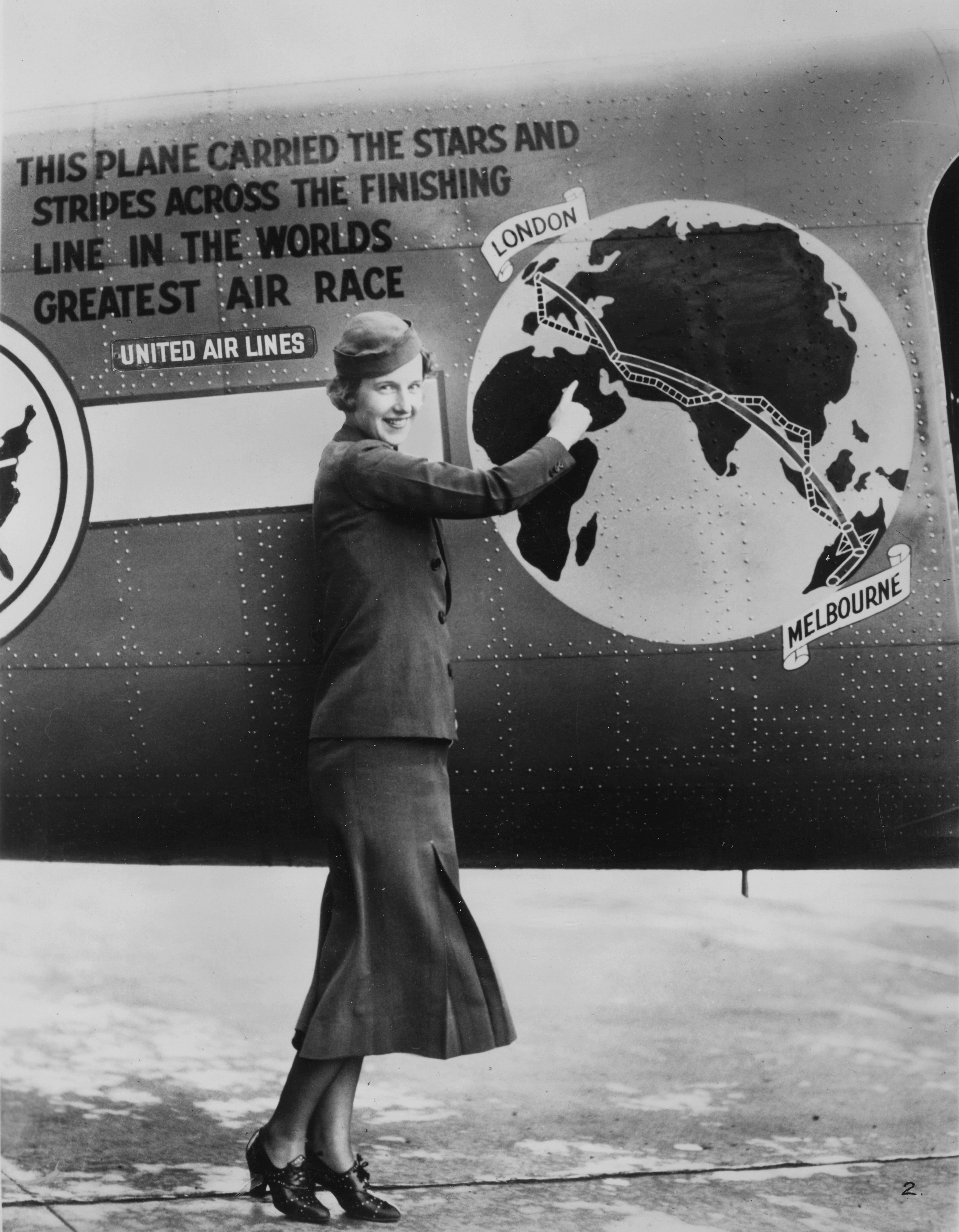 Black and white photograph of a woman in a stewardess uniform standing in front of a decorated airplane body. Plane body shows a globe with directional markers running from London to Melbourne.