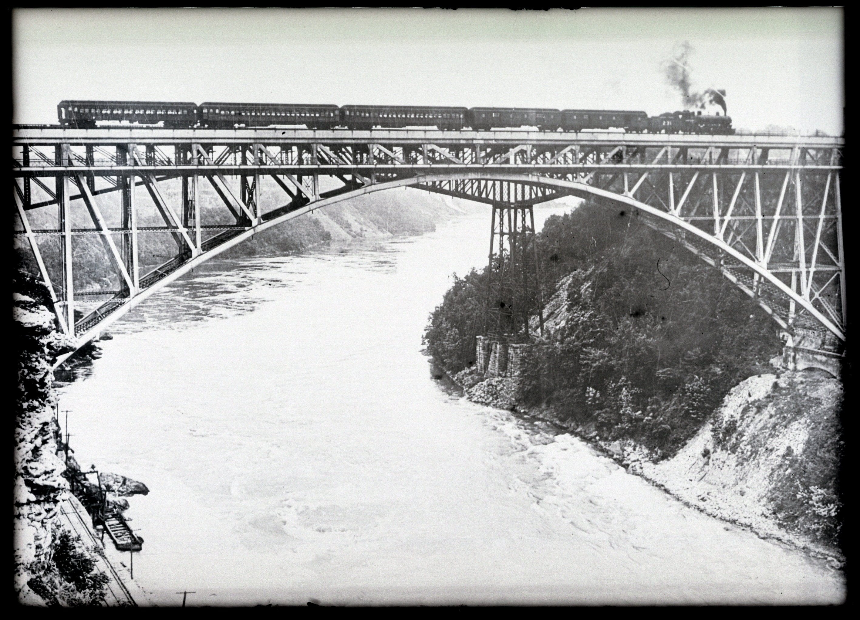 Black and white image of a train traveling on a steel arch bridge over the Niagara River rapids. 