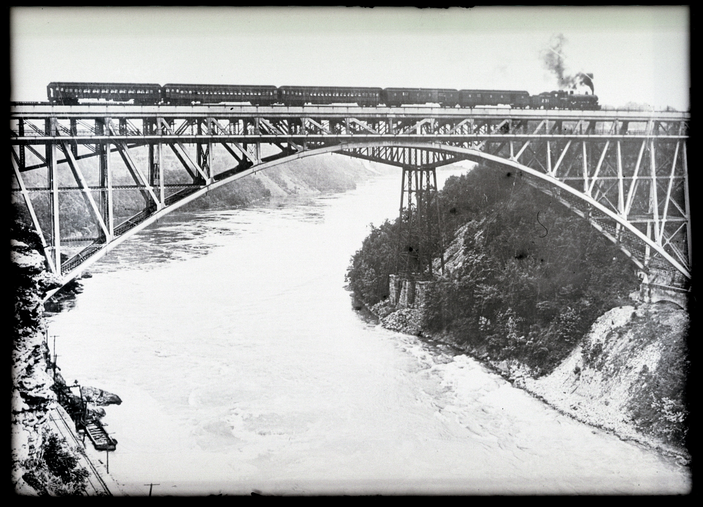 Black and white image of a train traveling on a steel arch bridge over the Niagara River rapids.