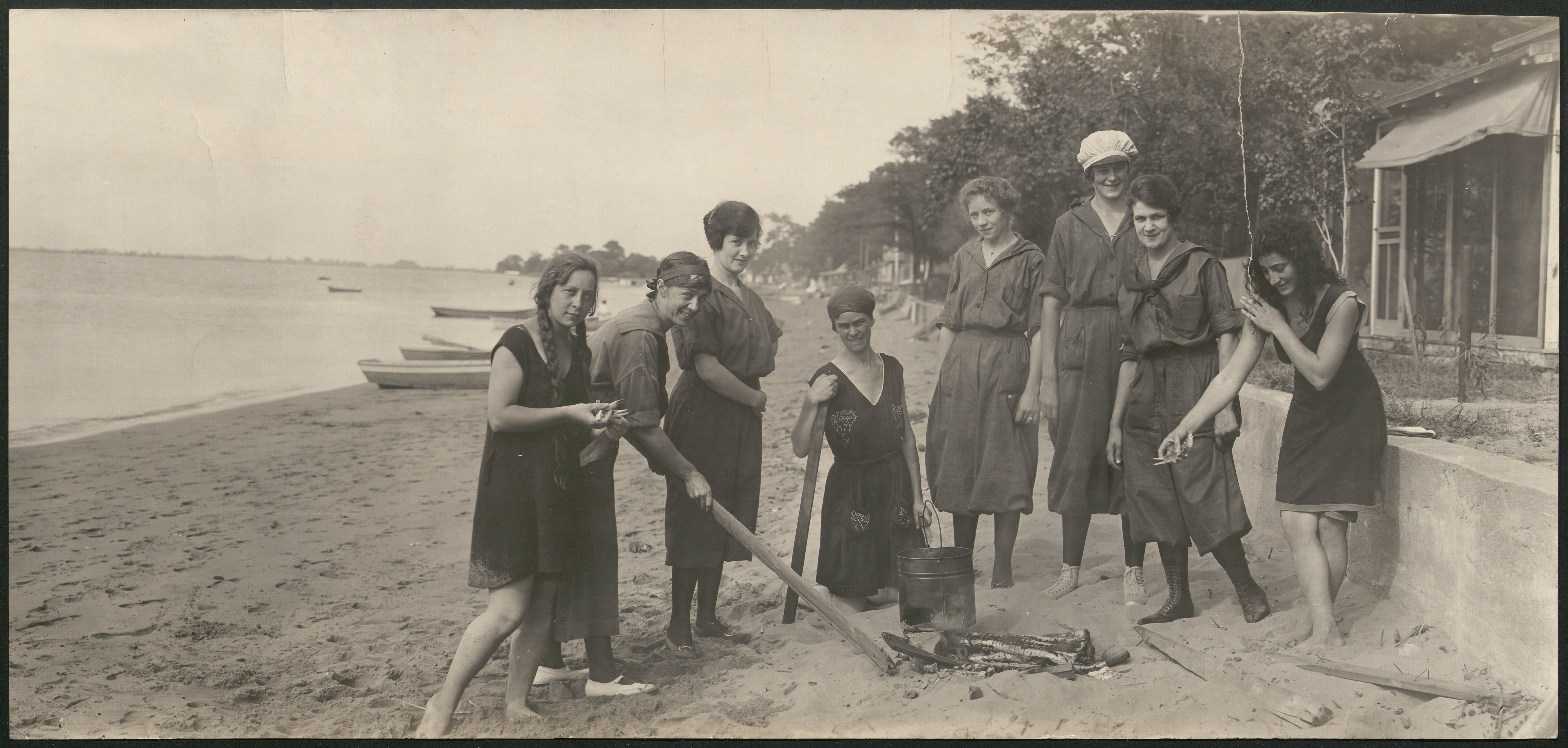 Panoramic black and white photograph of a group of women on a beach assembling a bonfire.