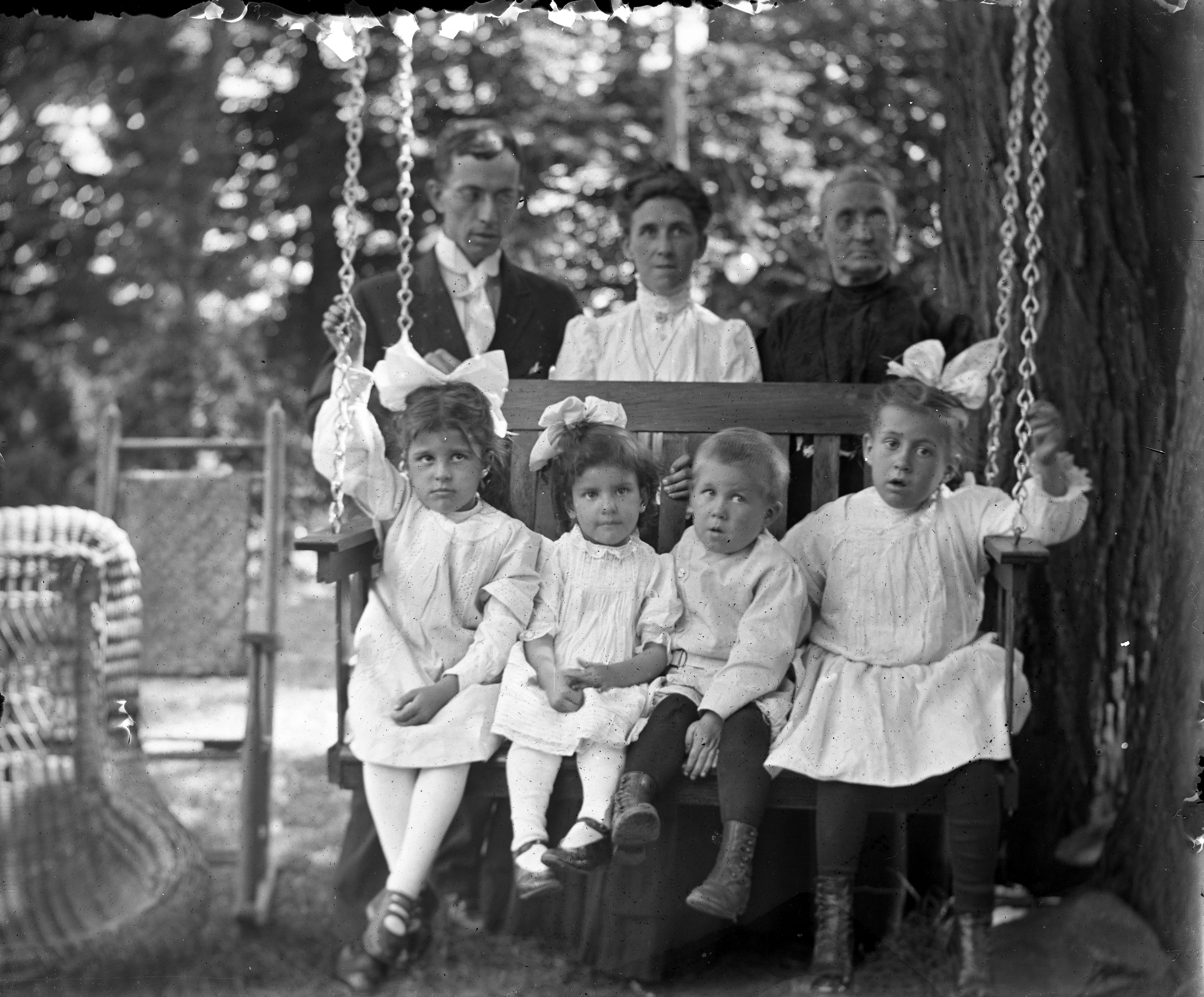 Black and white glass negative image of four children on an outdoor swing seat, with three adults behind them. 