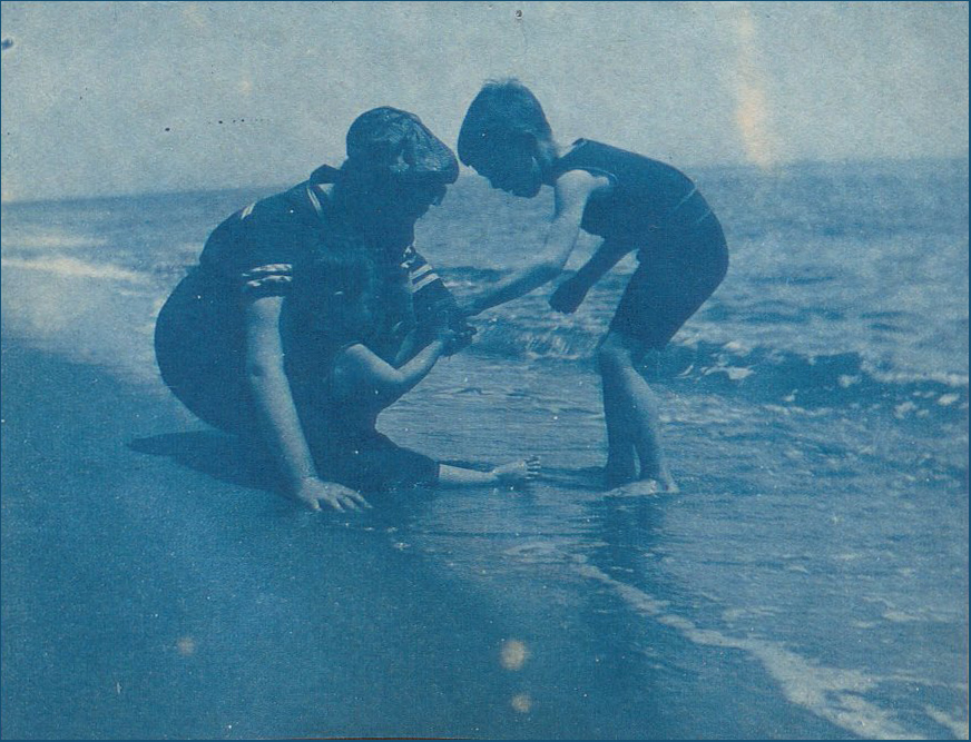 Cyanotype image of a person with two small children playing at the edge of the ocean.