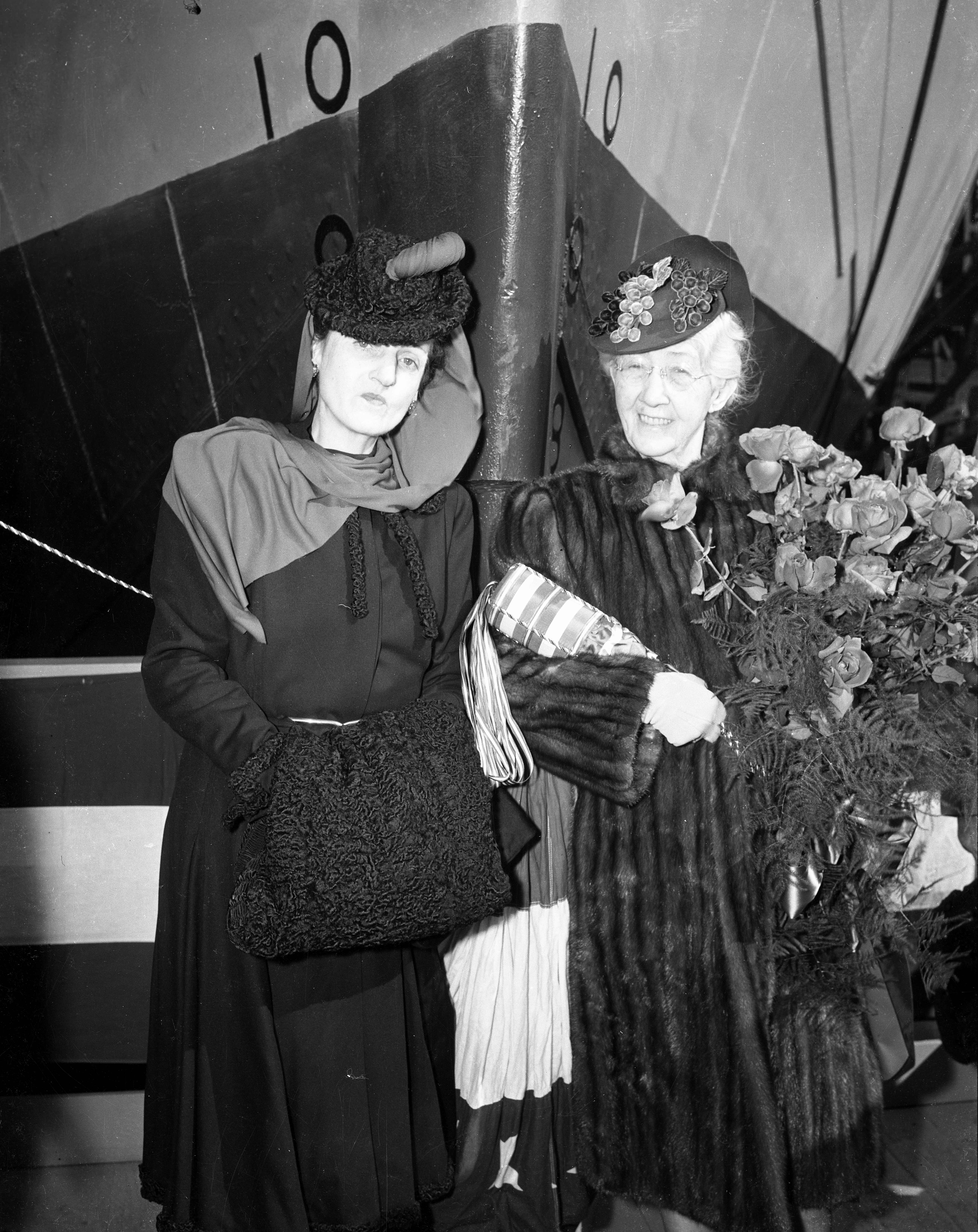 Posed portrait of two well-dressed women attending a ship launching. One is holding a ceremonial bottle and bouquet.