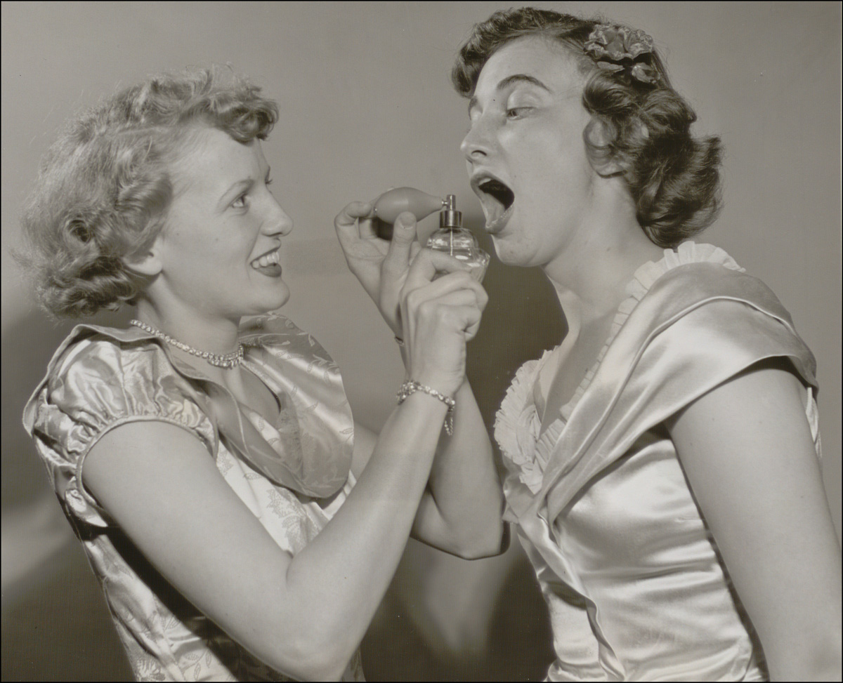 Black and white photo of two women. One is spraying something from a bottle into the other's mouth.