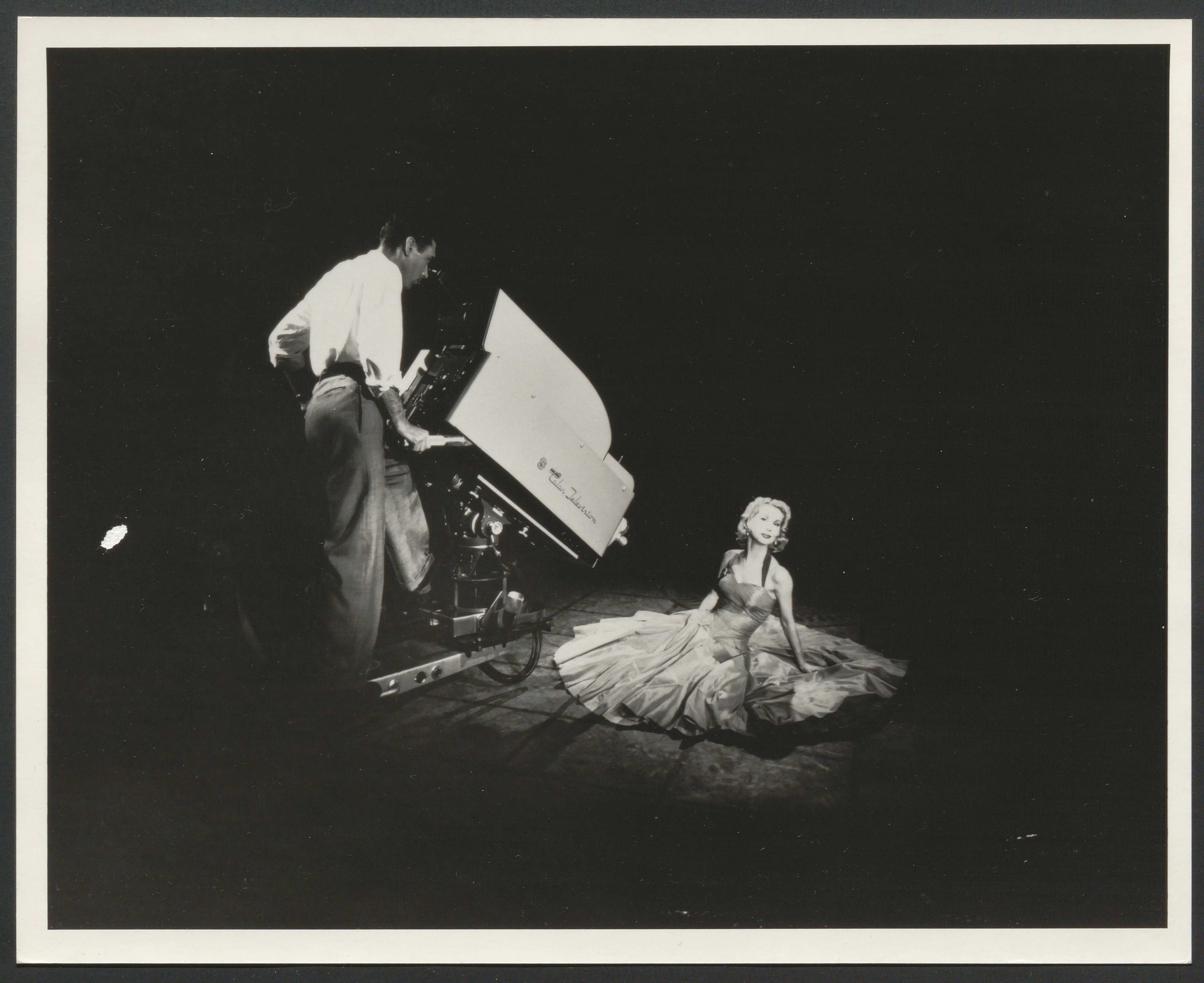 Black and white photograph showing a man aiming a very large television camera at a woman seated on the floor