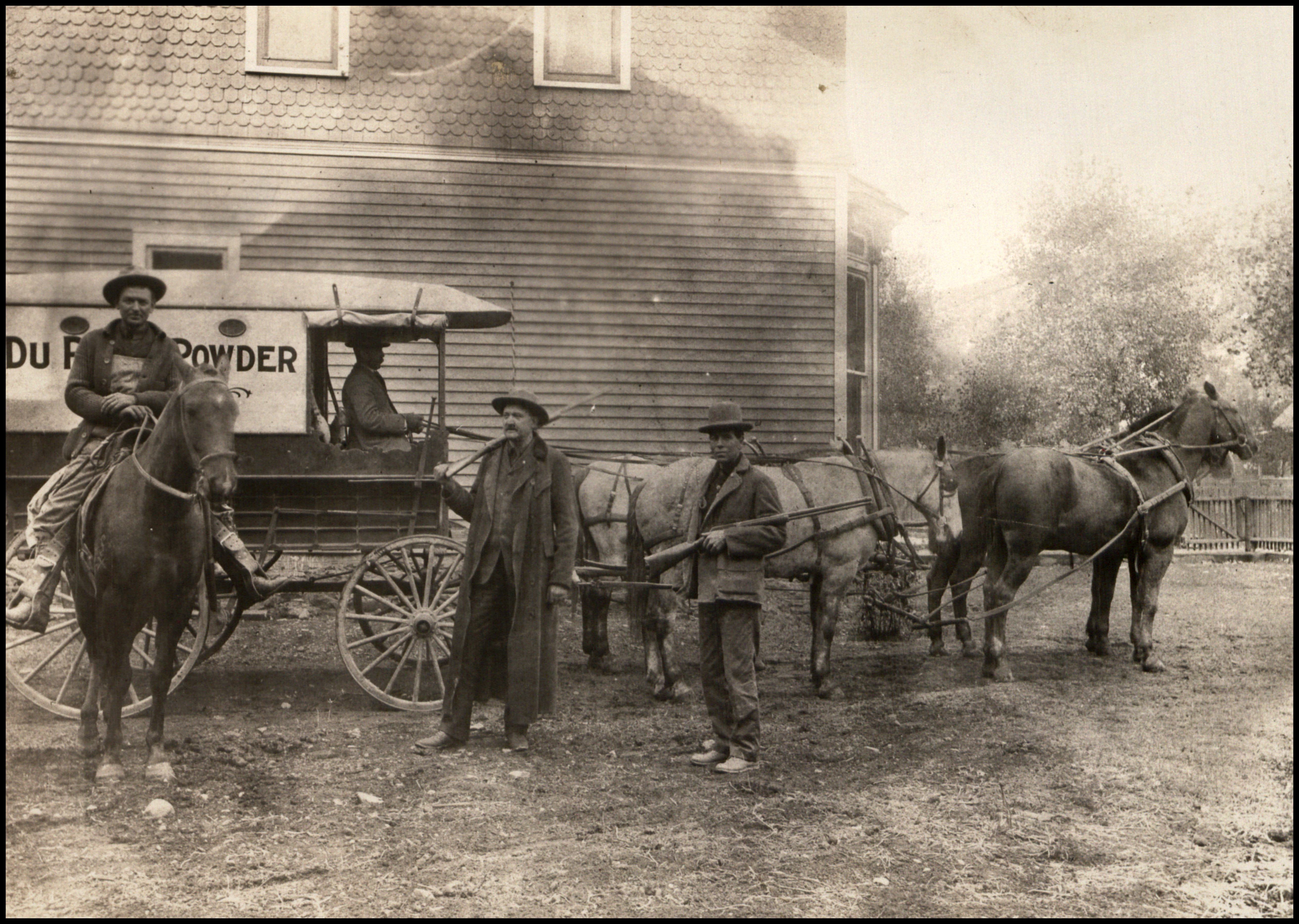 Black and white image of three men standing in front of a building and a horse-drawn wagon with 'Du Pont Powder' written on the side.