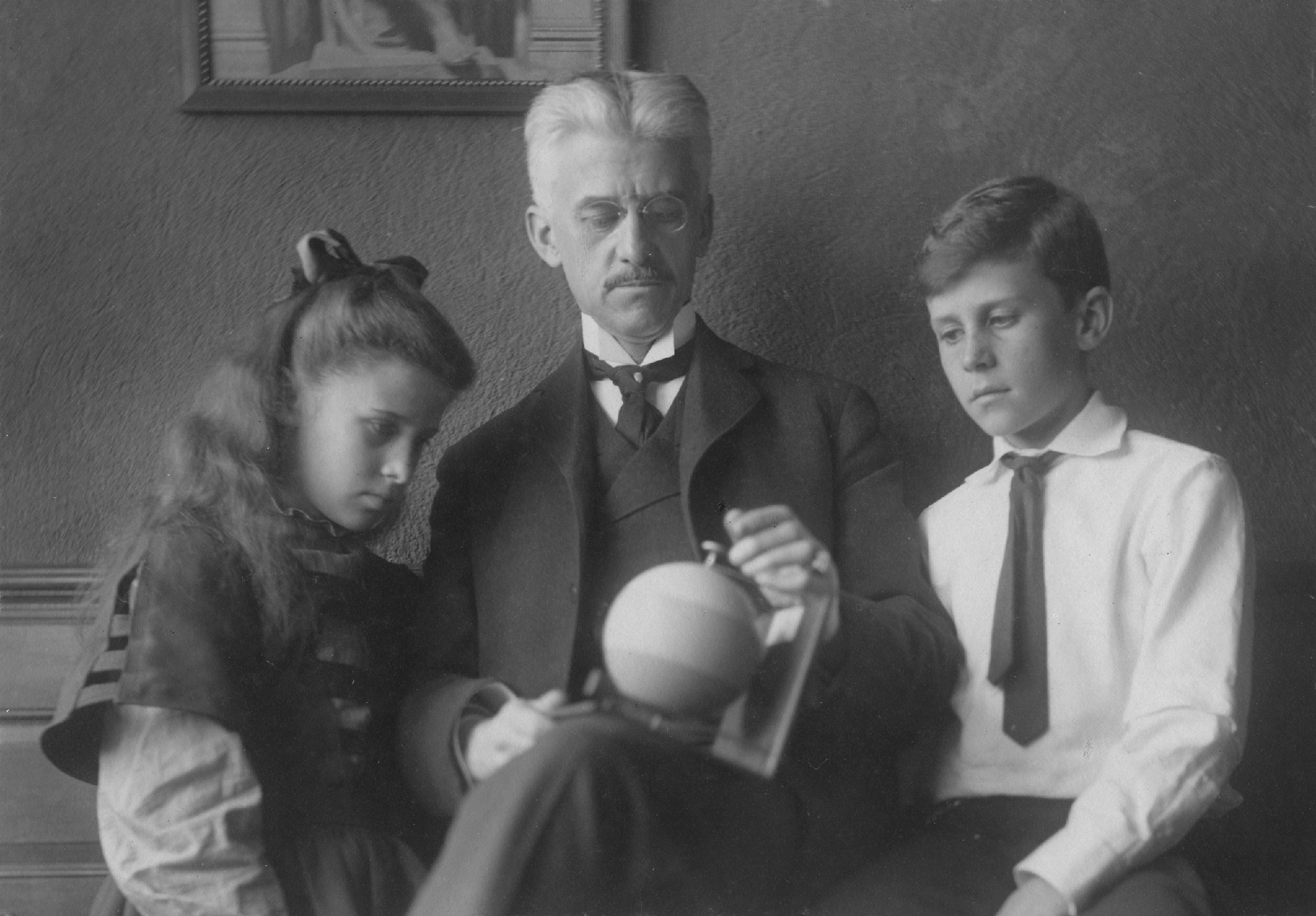 Black and white image of two children on either side of a man holding a striped sphere.