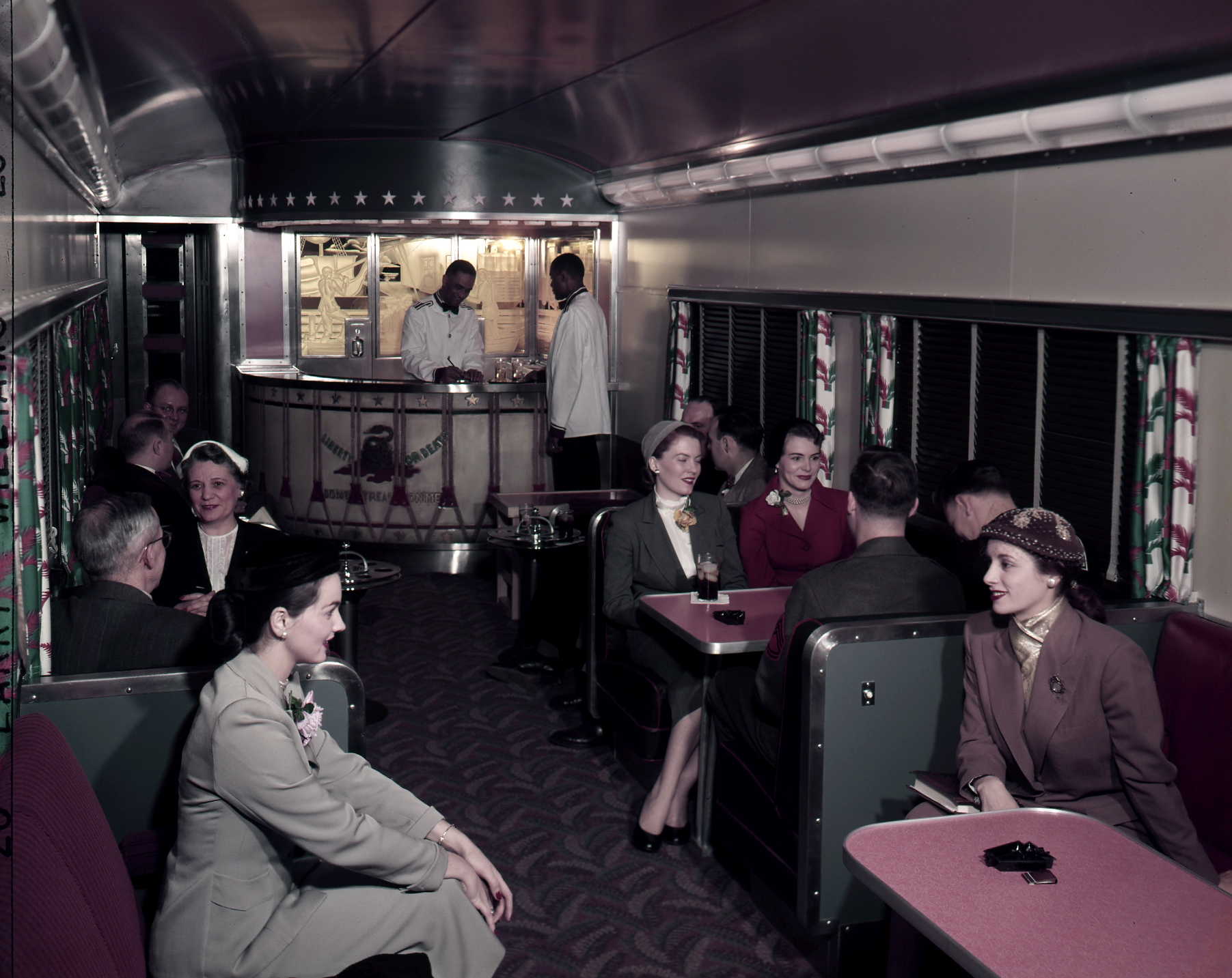 Color image of the interior of a dining car of a train, with passengers and staff.