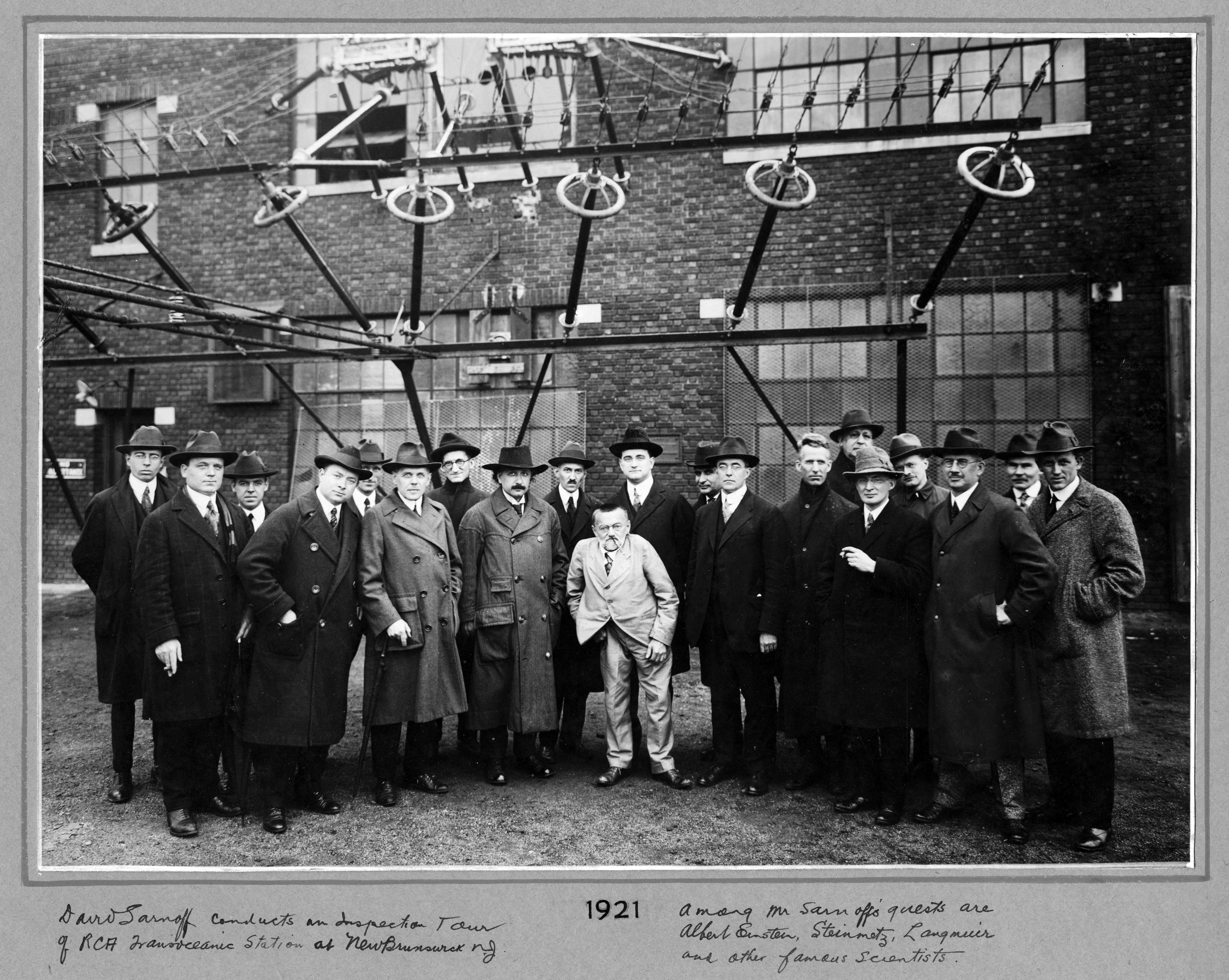 Black and white group photograph of men in suits and hats in front of an industrial building