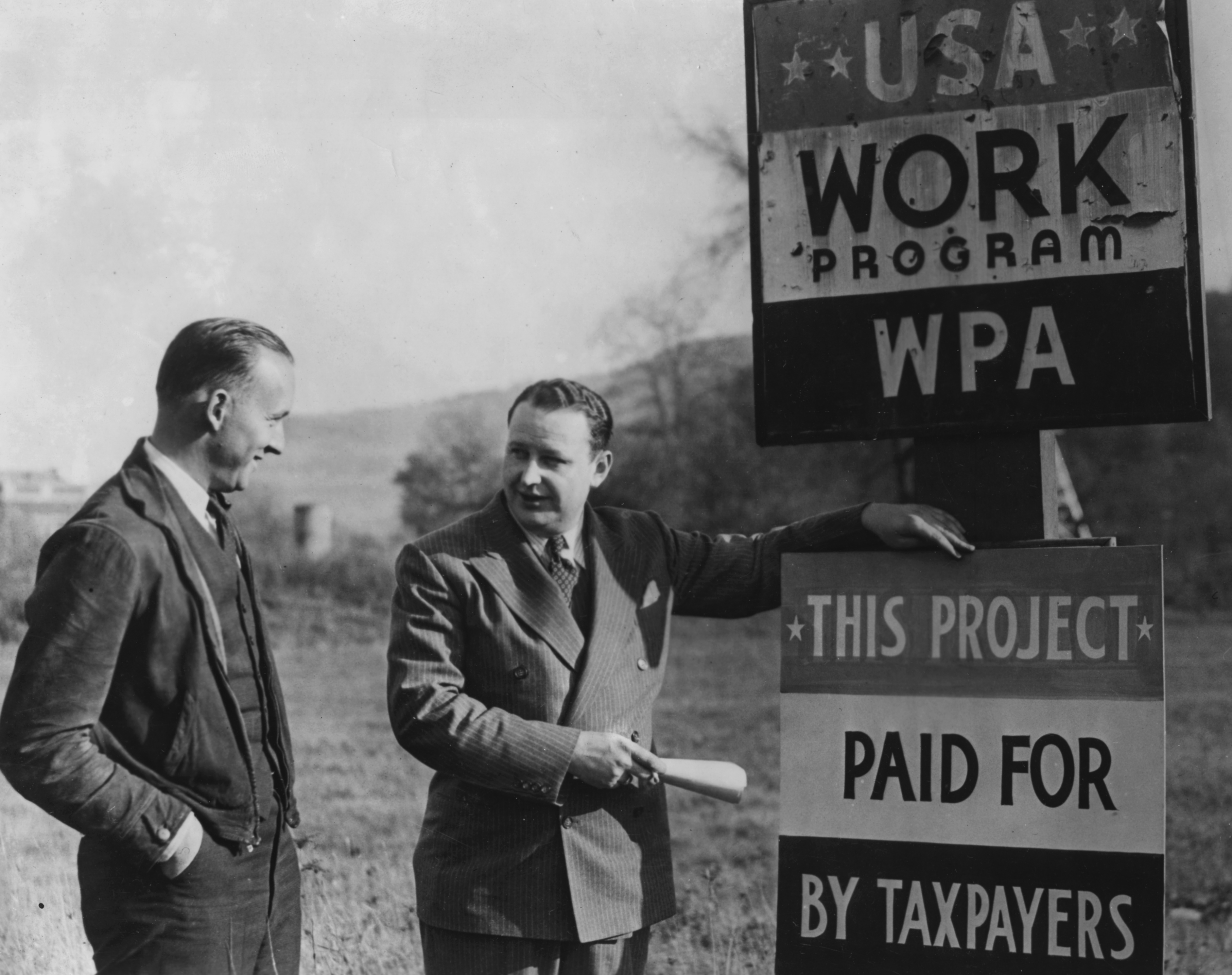 Two men standing near roadside signs declaring that roadwork is a WPA program paid for by taxes.